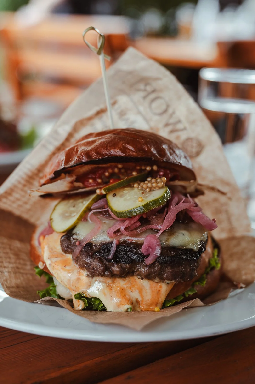 Ein Burger mit zwei Patties, gewürzt mit Senfsamen, Zwiebeln, Gurkenscheiben, Salat, und Käse, serviert auf braunem Papier in einem Restaurant.