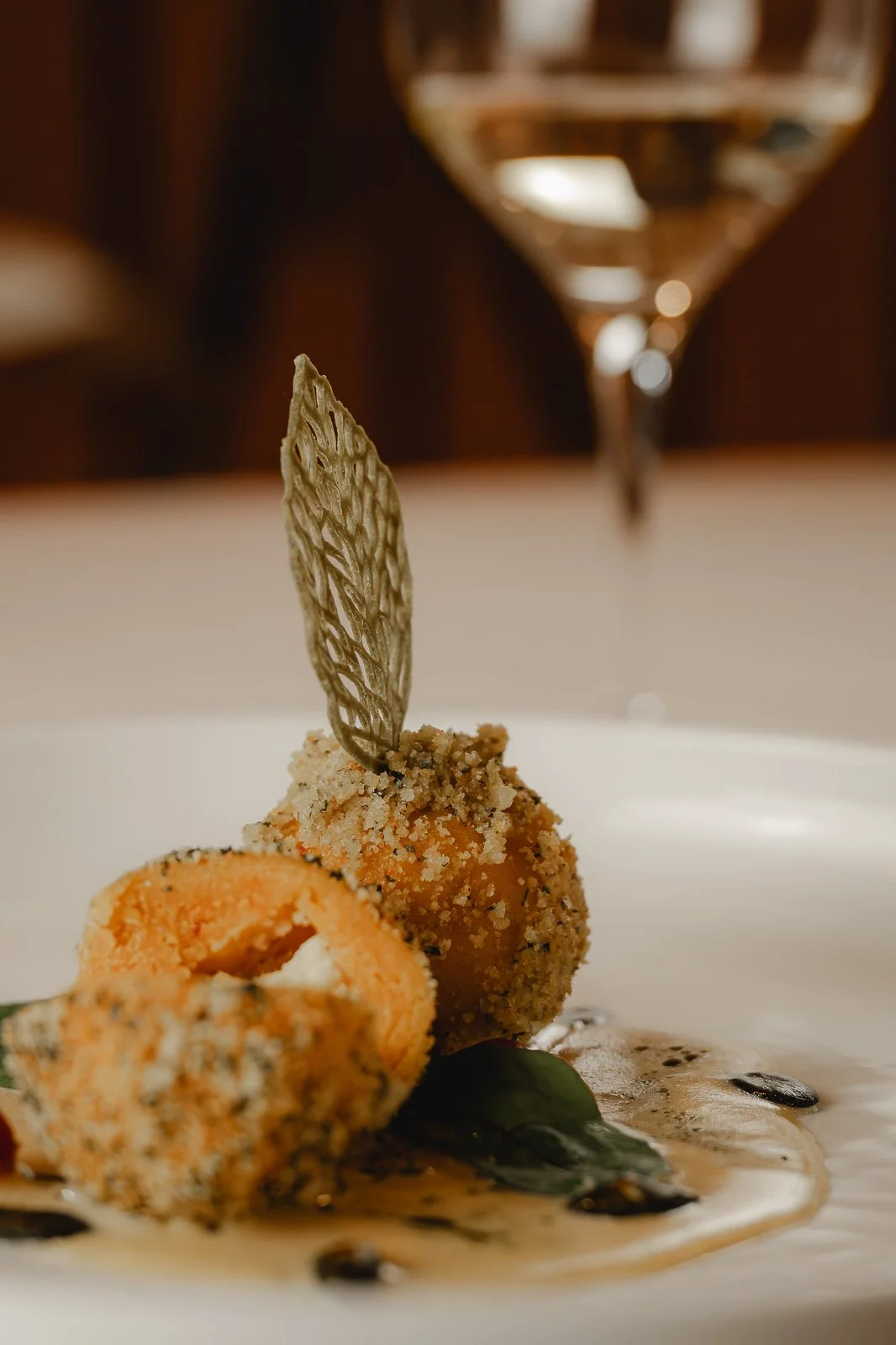 Frittierte Meeresfrüchte mit einer dekorativen Spiegelei-Teile auf einem weißen Teller, garniert mit einer hauchdünnen, goldenen essbaren Dekoration und einer grünen Blattform, serviert mit einer dunklen Sauce und einer Weinglas im Hintergrund. 