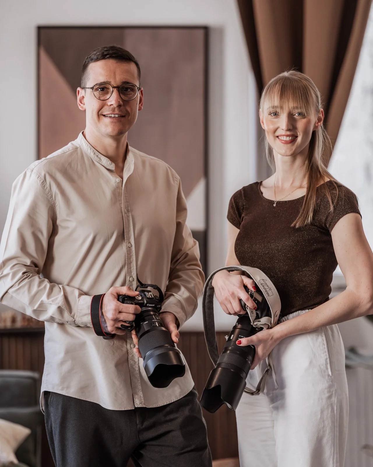 Ein Mann und eine Frau stehen in einem Raum und halten Fotografien. Beide tragen Kameras, der Mann trägt eine beige Bluse und die Frau ein dunkles Oberteil. Sie sind beide lächelnd und schauen zur Kamera.