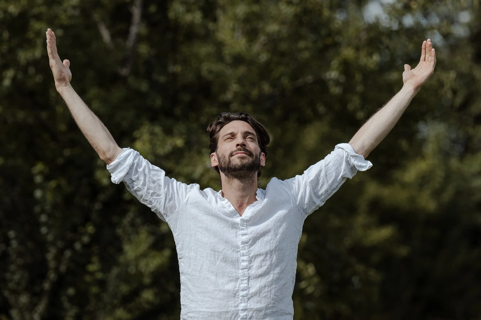 Ein Mann mit geschlossenen Augen und einem weißen Hemd steht im Freien mit ausgebreiteten Armen, umgeben von Bäumen.