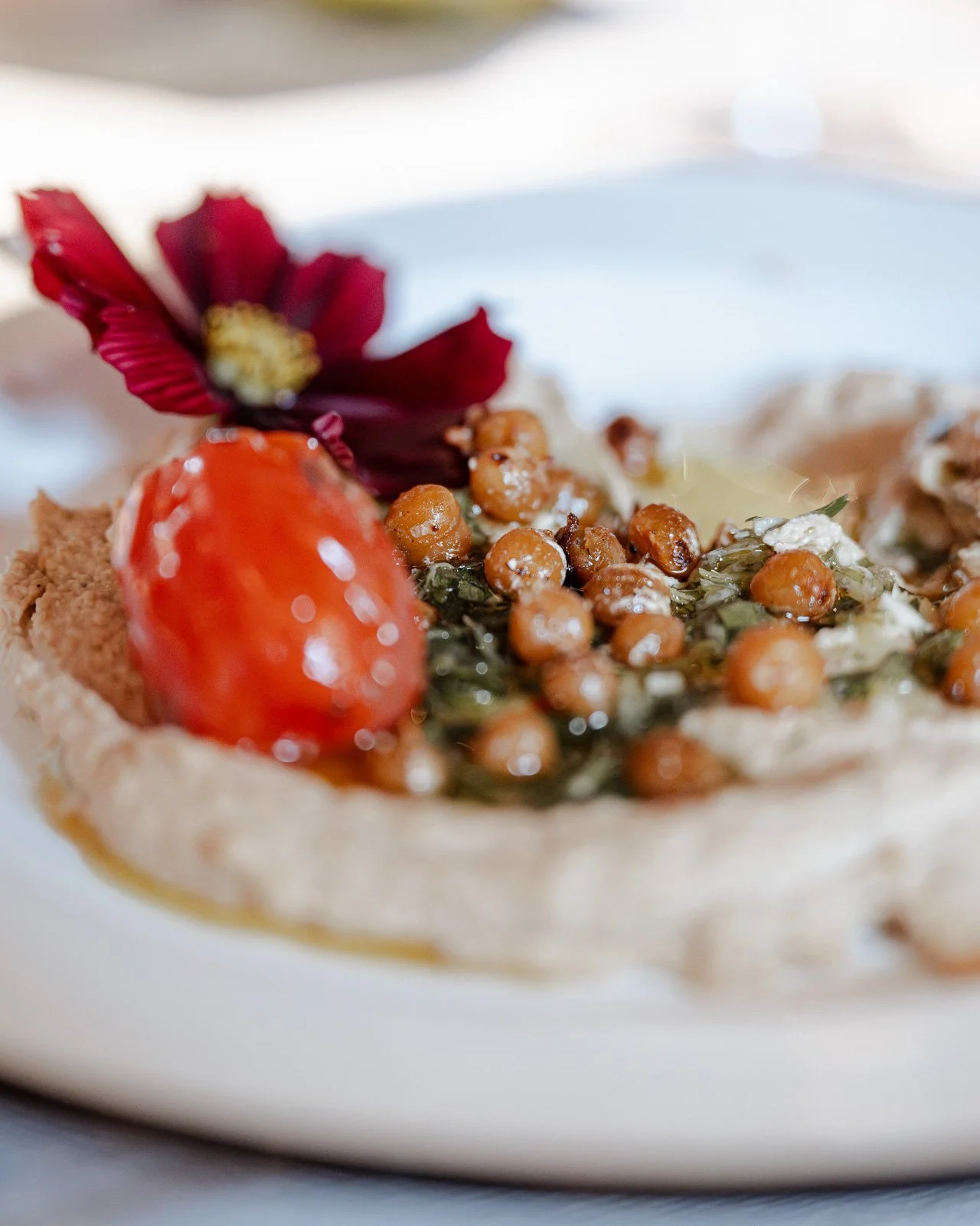 Nahaufnahme eines hellen Tellers mit Hummus, garniert mit Tomaten, Kräutern und einem Blütenblatt.
