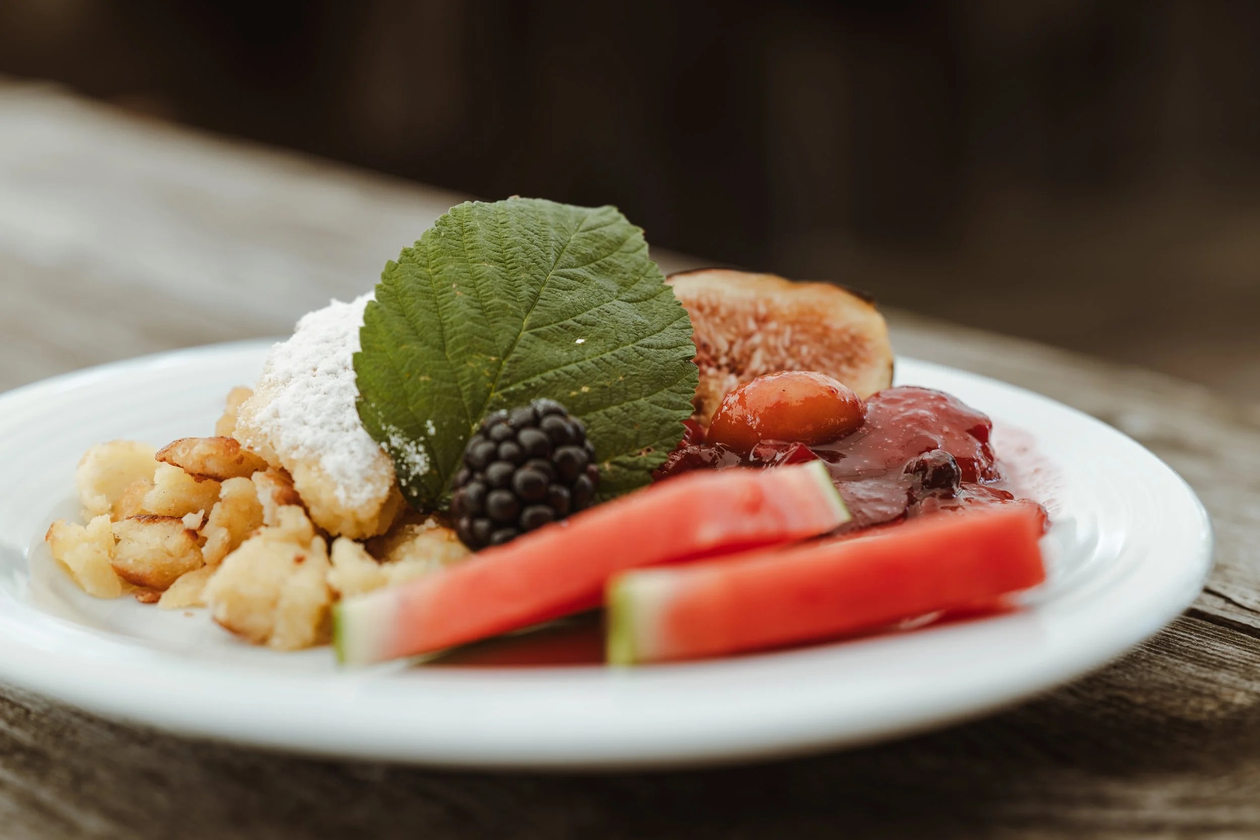 Ein Teller mit Obst und Süßspeisen, darunter Wassermelone, Feigen, Beeren, eine große grüne Weinblatt, Streusel und Marmelade.