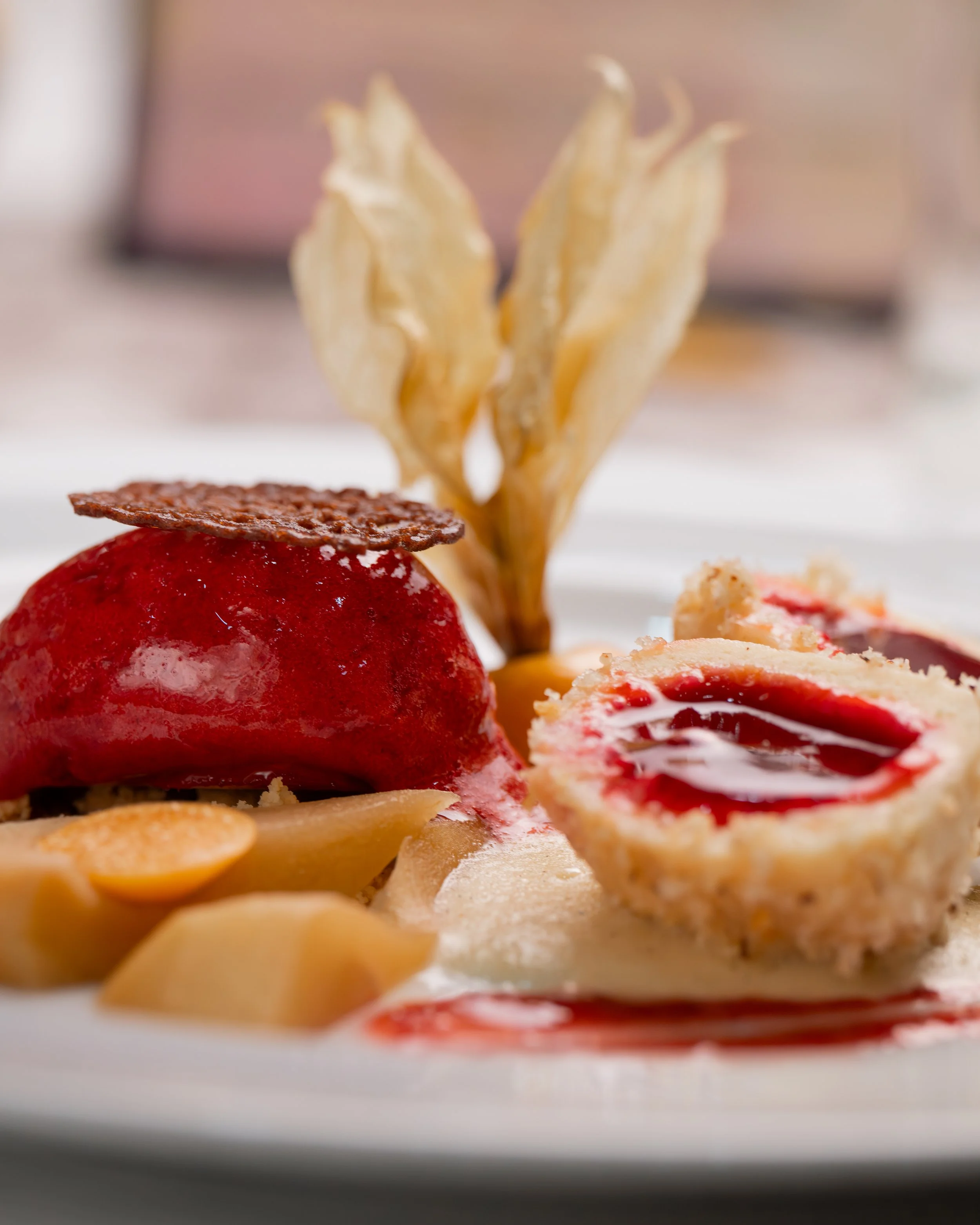 Ein Teller mit einem roten Fruchtdessert, einer goldenen Sauce, knusprigem Gebäck um die Frucht, einer dünnen Keks- oder Schokoladentafel, einer Knusperspirale und weiteren Fruchtstücken