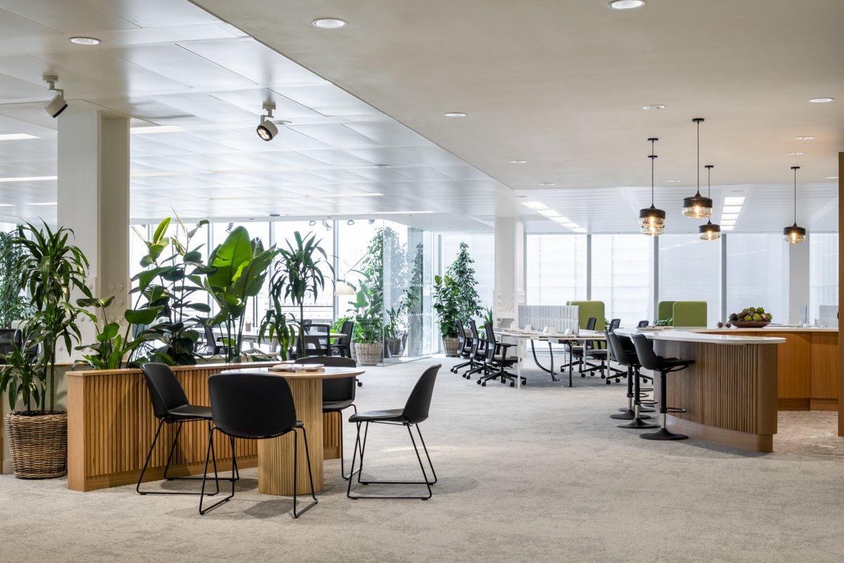Modern office space with multiple tables and chairs, green plants, large windows, and a kitchen area with bar stools and pendant lighting.