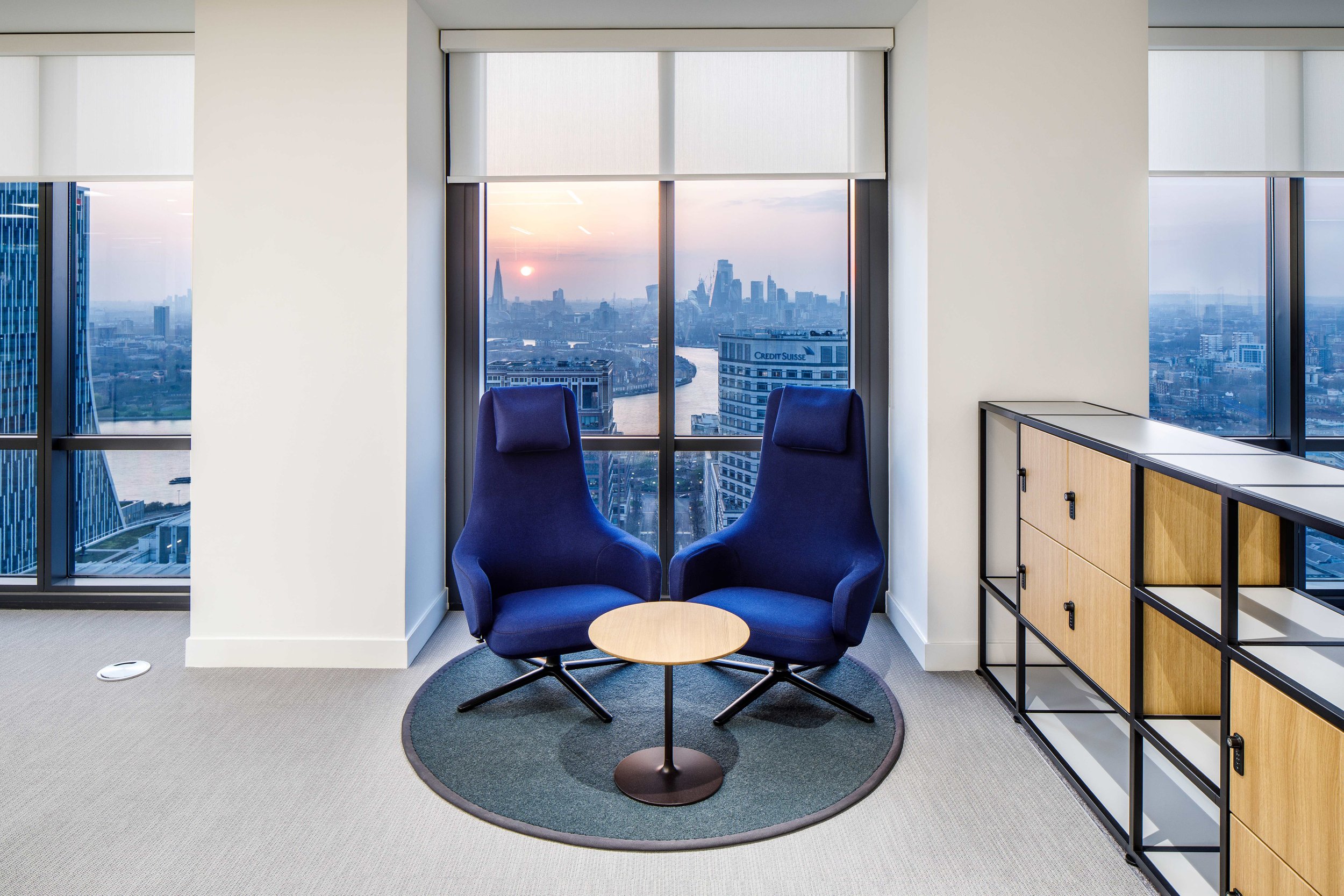 Modern office space with two blue high-backed chairs and a small round wooden table, facing large glass windows showcasing a cityscape at sunset with a river, various buildings, and a partly cloudy sky.