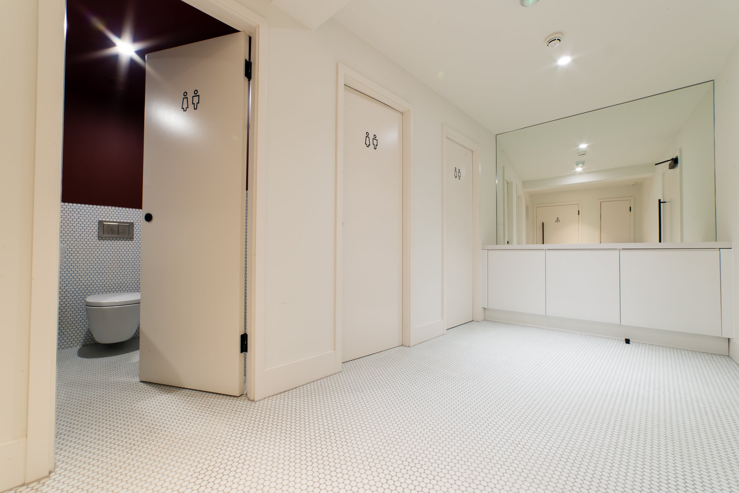 A clean, white public restroom with a large mirror, multiple doors with gender signs, and a toilet visible through the open door.