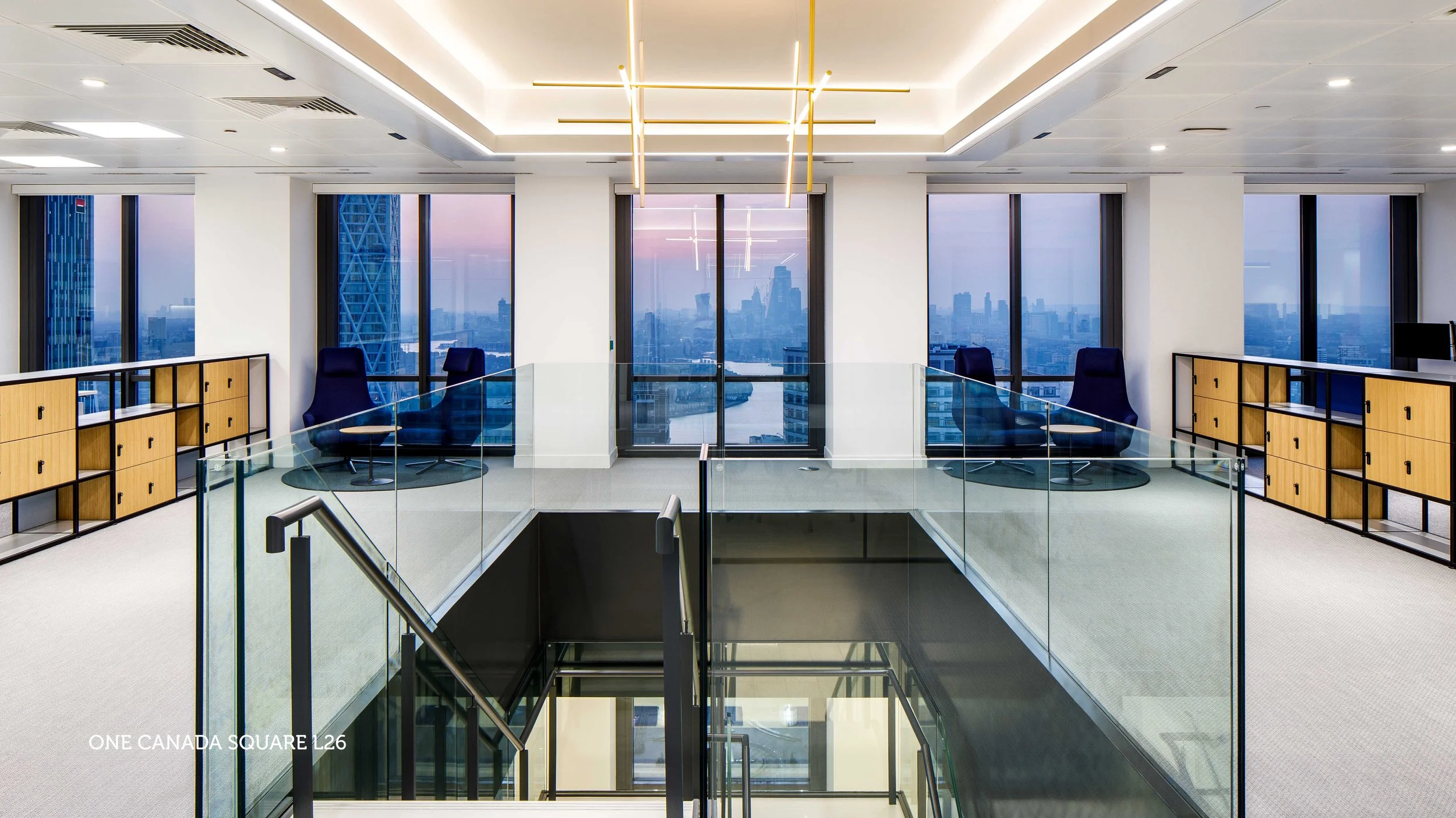 Modern high-rise office with large floor-to-ceiling windows, a blue sofa, a white lounge chair, a glass coffee table, a beige rug, and a contemporary ceiling light fixture.
