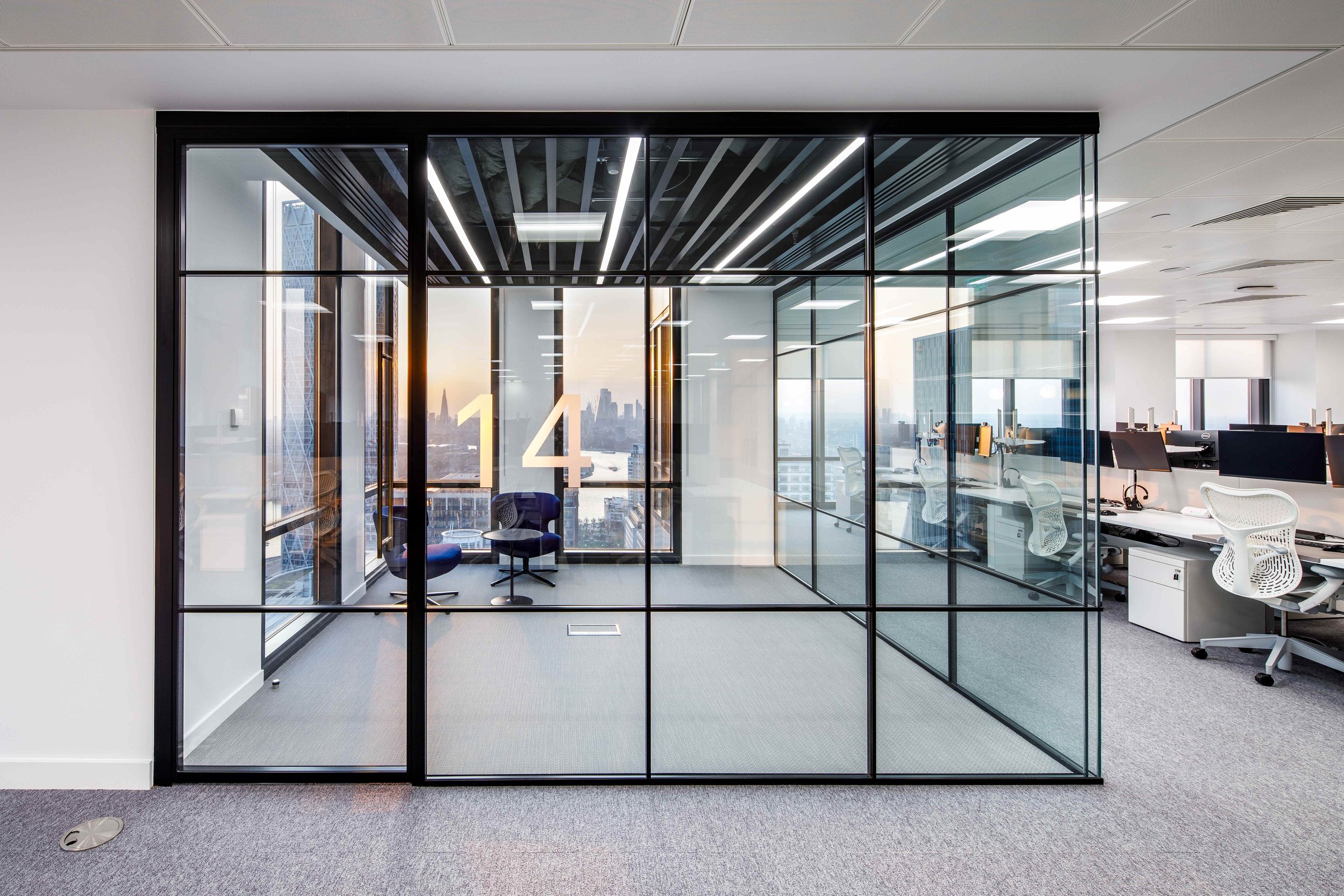 Modern office space with glass walls, city skyline view, and seating area with two blue chairs and a small round table.