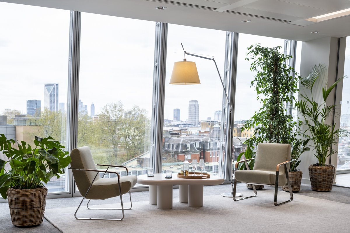Modern office lounge area with two beige armchairs, a round coffee table with glasses, a floor lamp, and potted green plants in front of large windows with a city skyline view.