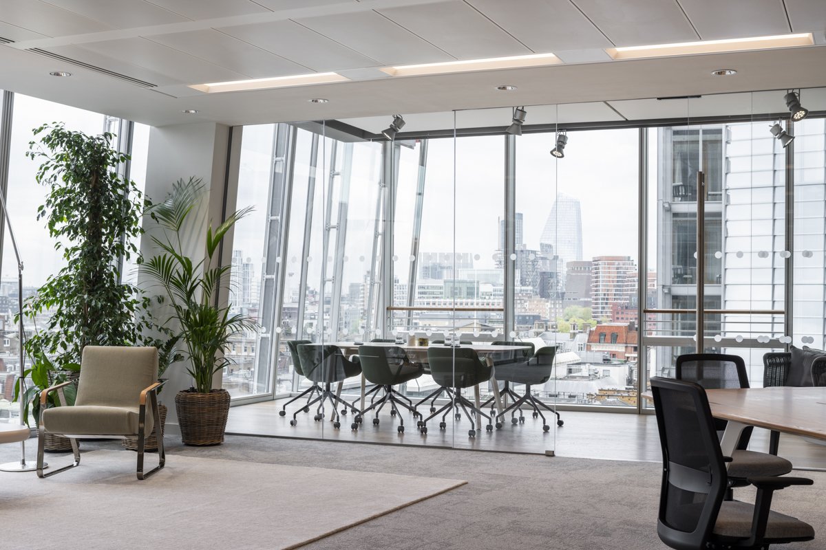 Modern office conference room with large glass windows, round table, and black chairs, overlooking city skyline with tall buildings and skyscrapers, daytime.