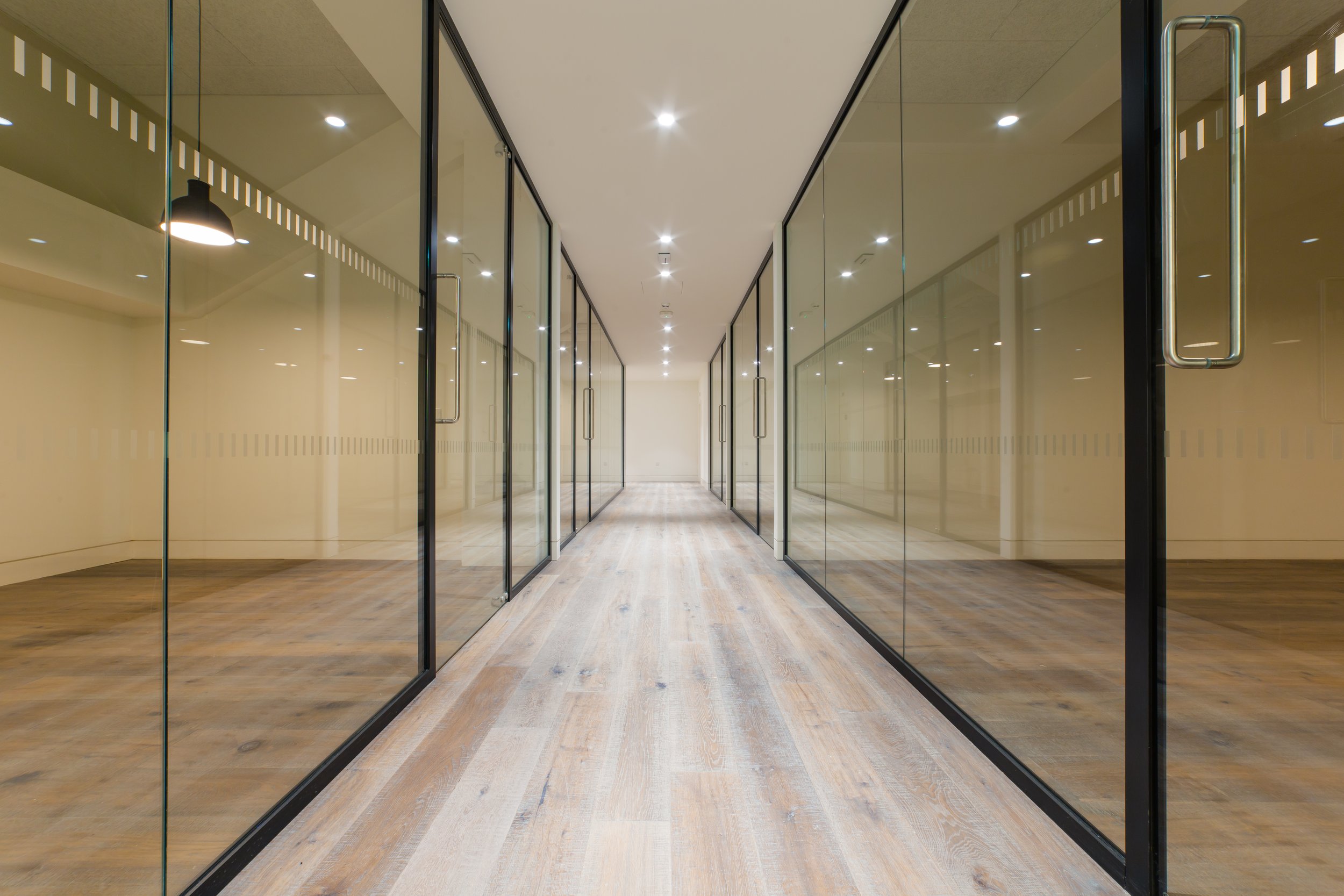 Empty modern office corridor with glass-walled rooms on either side and wooden floors.