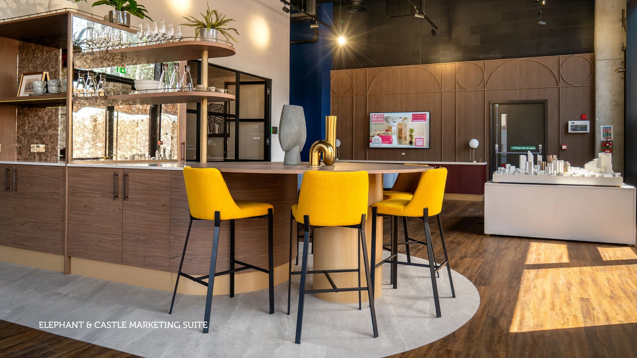 Modern cafe lounge with pink armchairs, black circular coffee table, checkered rug, and a well-lit cafe counter in the background.