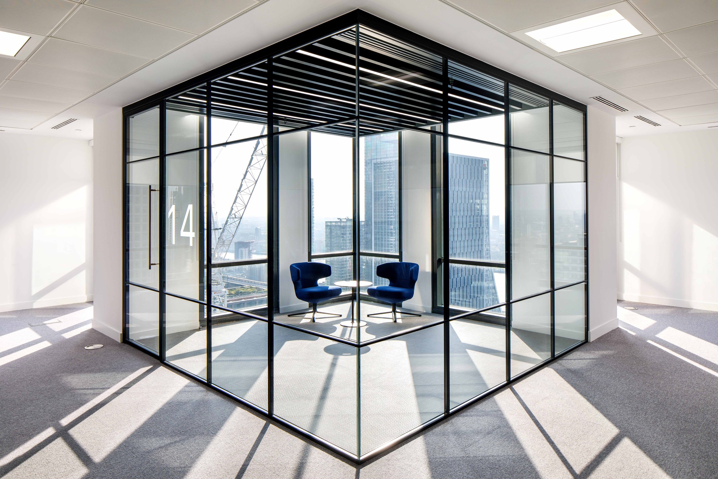 Modern office corner with two blue chairs and a small table inside a glass enclosure, overlooking a city skyline with tall buildings and construction cranes.