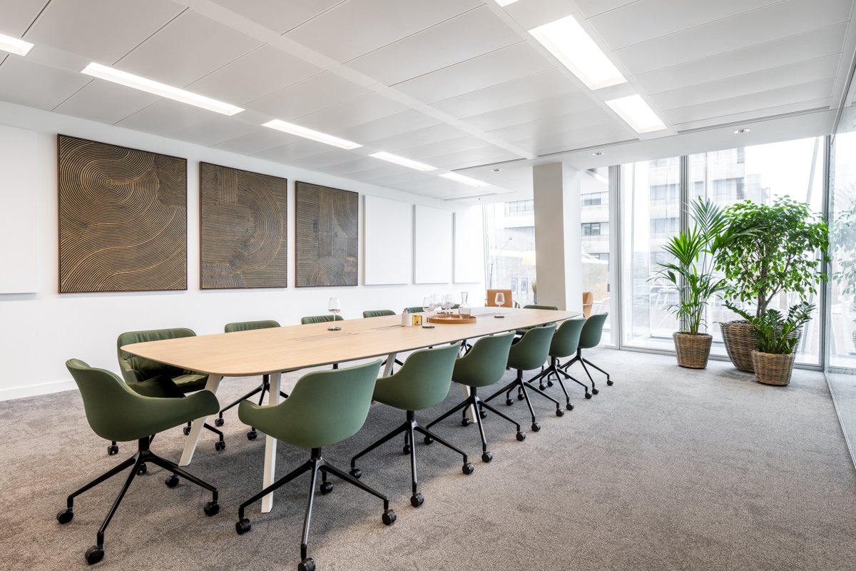 Modern conference room with a large wooden table, green chairs, abstract wall art, potted plants near floor-to-ceiling windows, and bright ceiling lights.