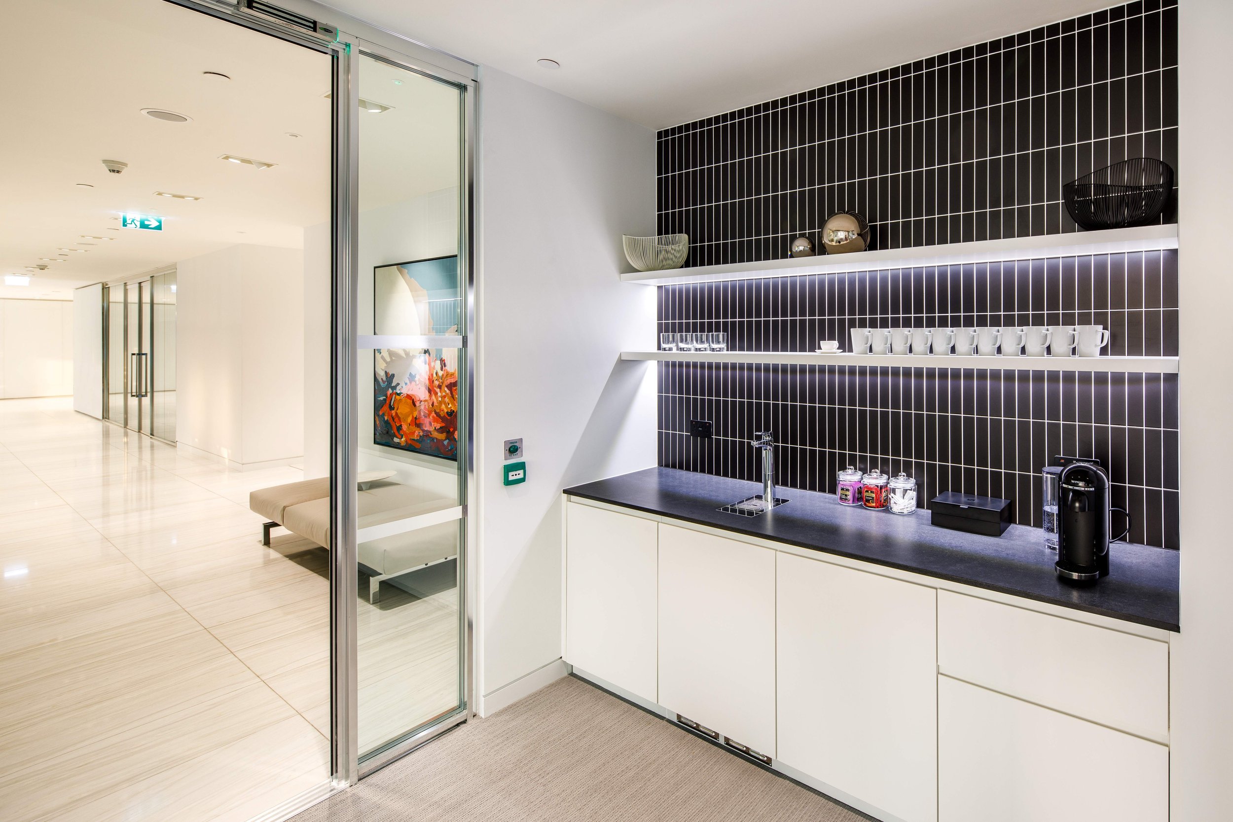 Modern kitchenette with black tiled backsplash, white cabinets, small sink, coffee machine, glass jars, and open shelves with cups and decorative items, adjacent to a hallway with art and seating area.