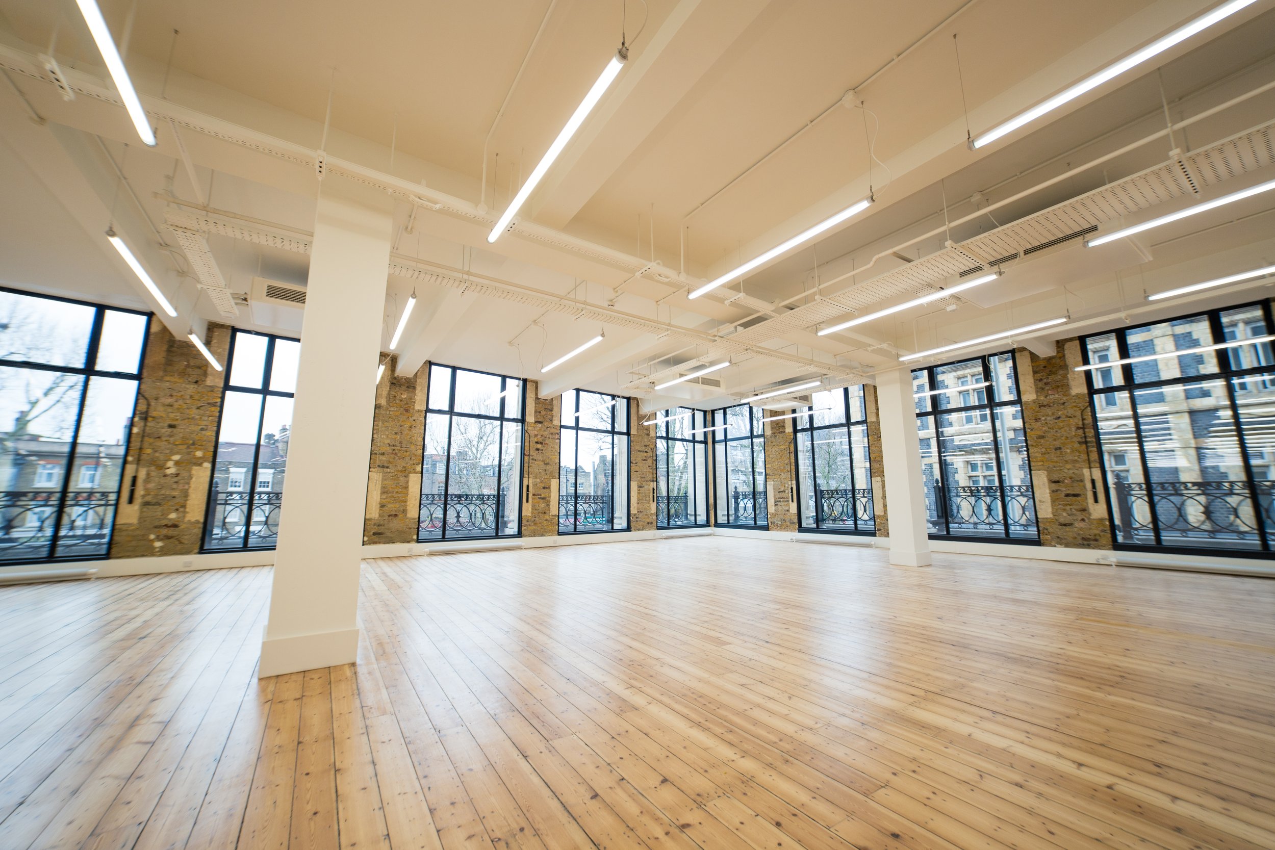 Empty bright loft with large windows, wooden floors, exposed brick walls, and ceiling lights.