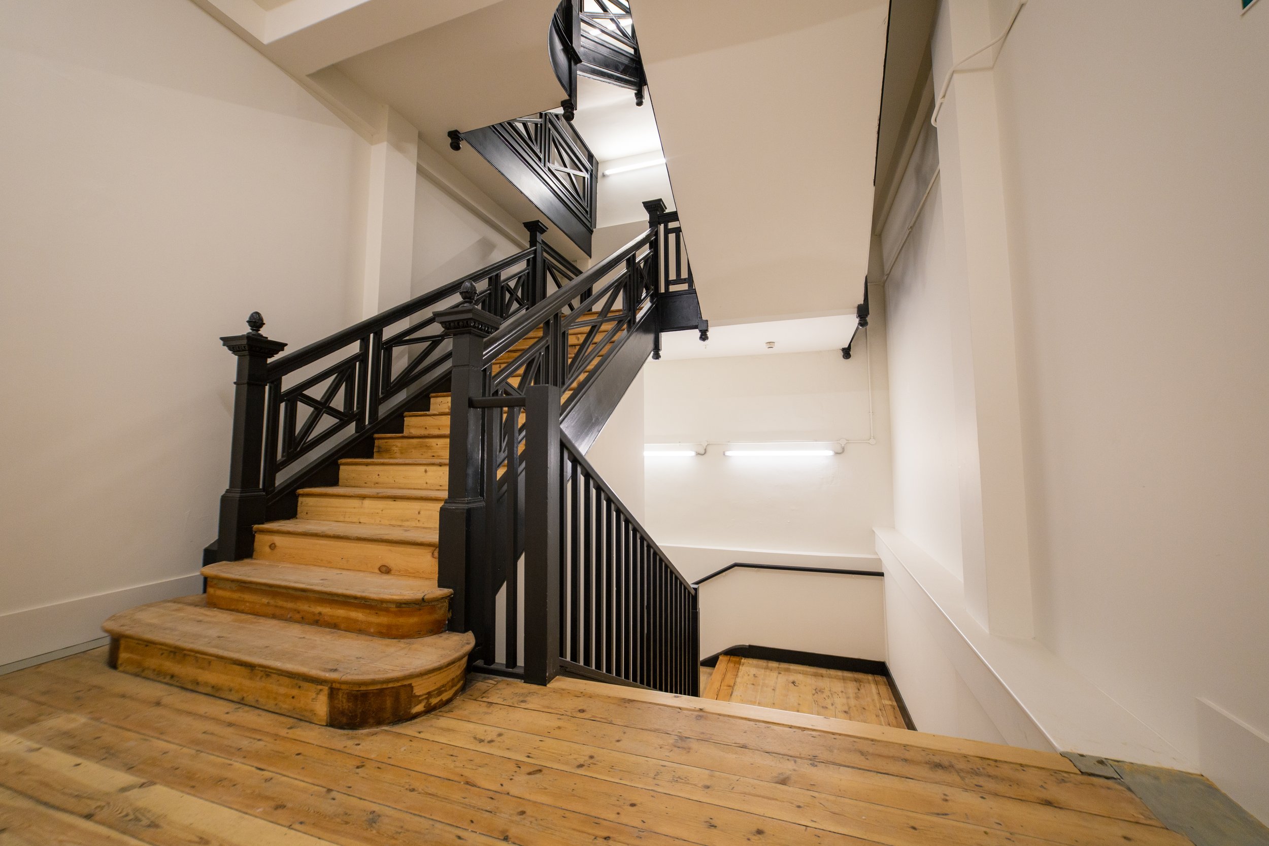 Interior view of a staircase with wooden steps and a black metal railing, in a well-lit building with white walls and a light-colored wooden floor.