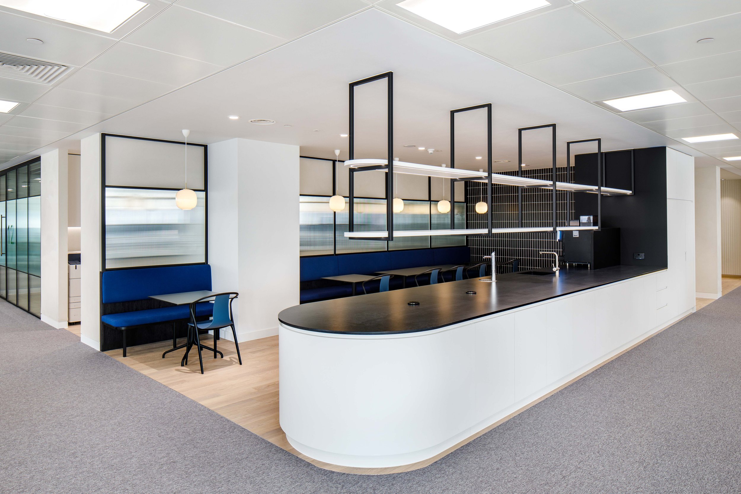 Modern office break area with a curved white counter, black countertop, and blue seating along the wall. Pendant lights and a black shelving structure hang above a small countertop area.