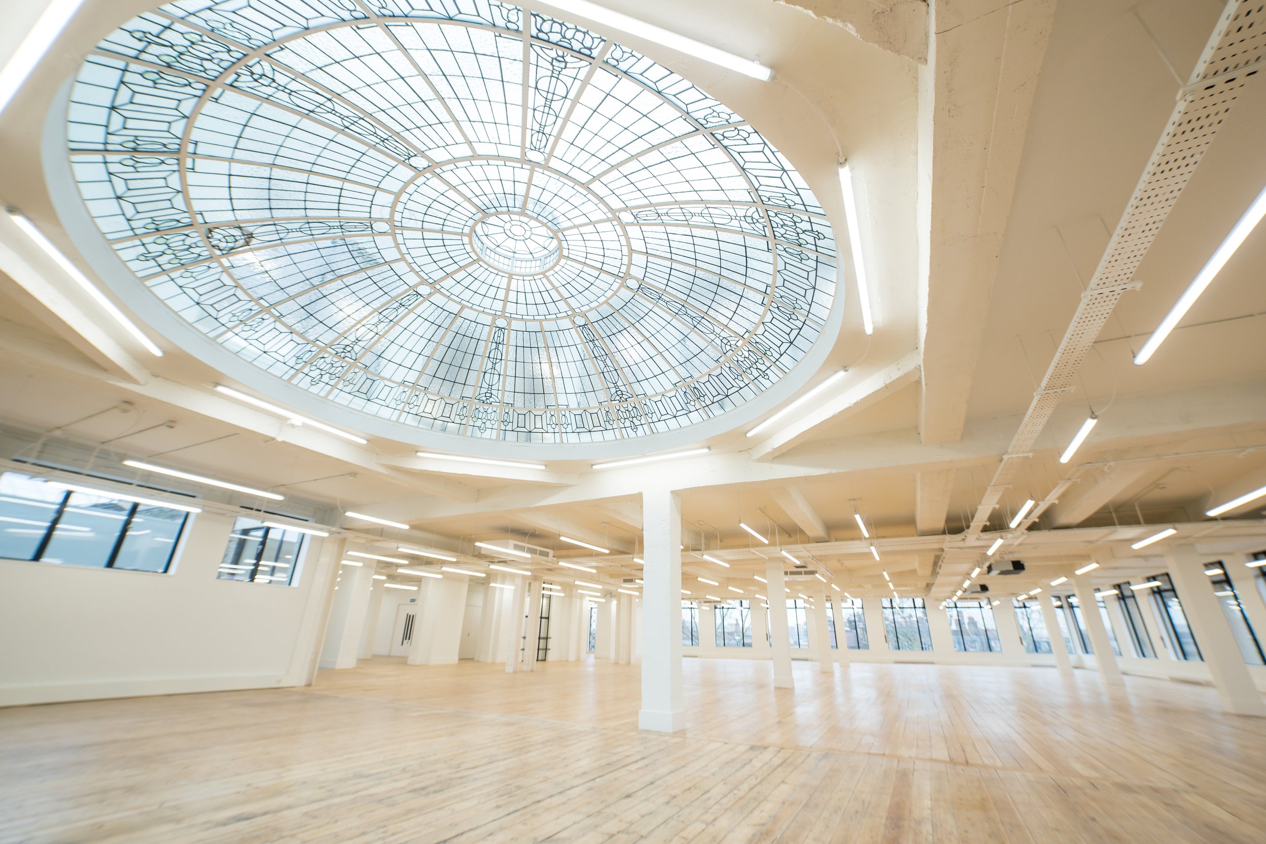 Interior view of an empty room with wooden floors, white walls, large windows, and a decorative stained glass ceiling.