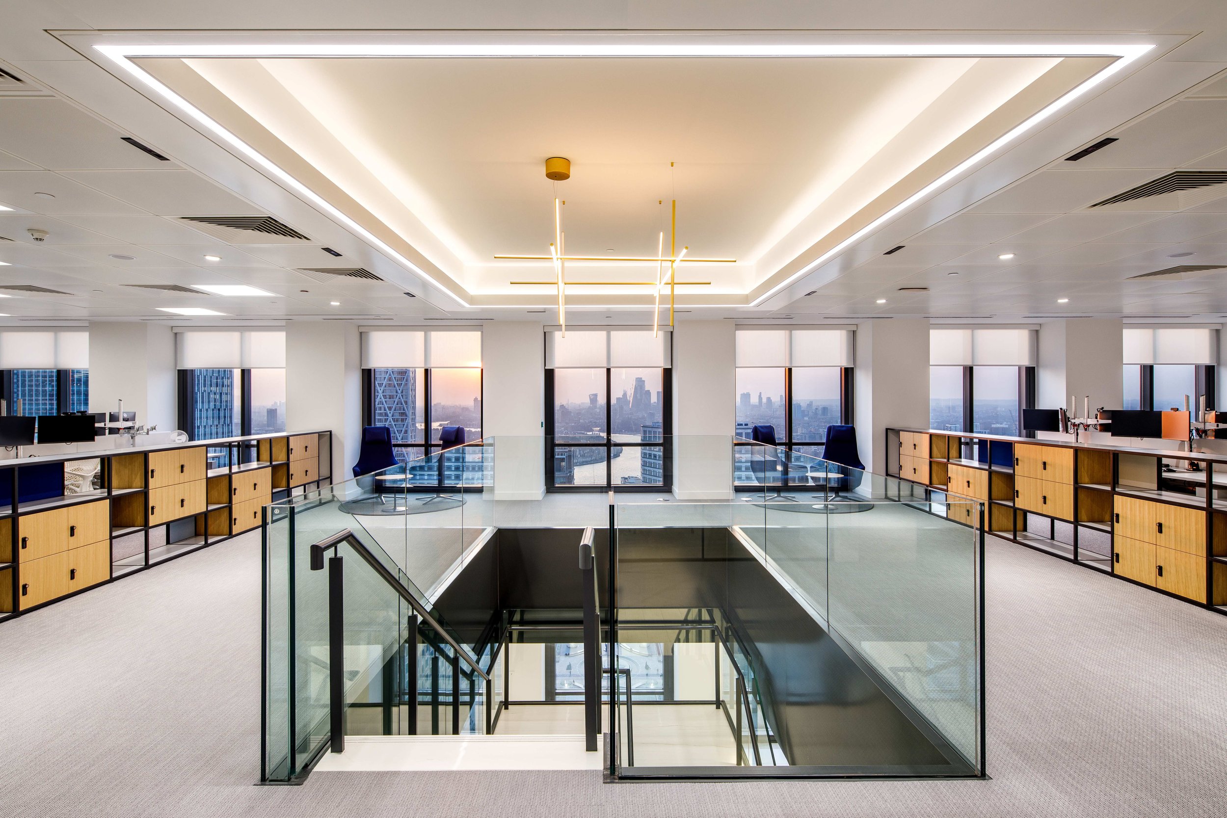 Modern office space with large windows, city skyline view, glass railing around open staircase, fluorescent and pendant lighting, and rows of wooden cabinets and workstations.