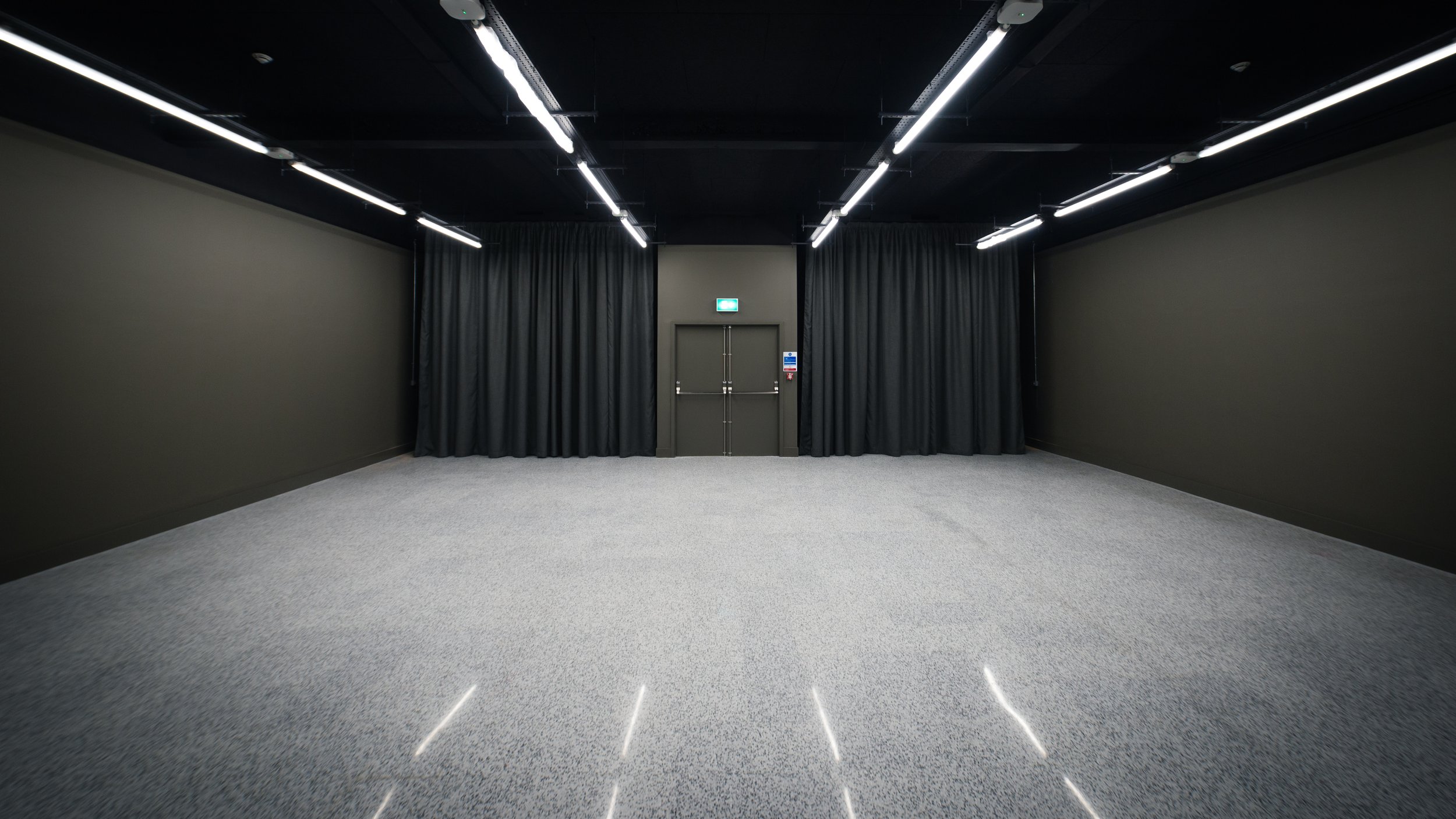 Empty room with black walls, black curtains, a grey door, and ceiling with strip lighting