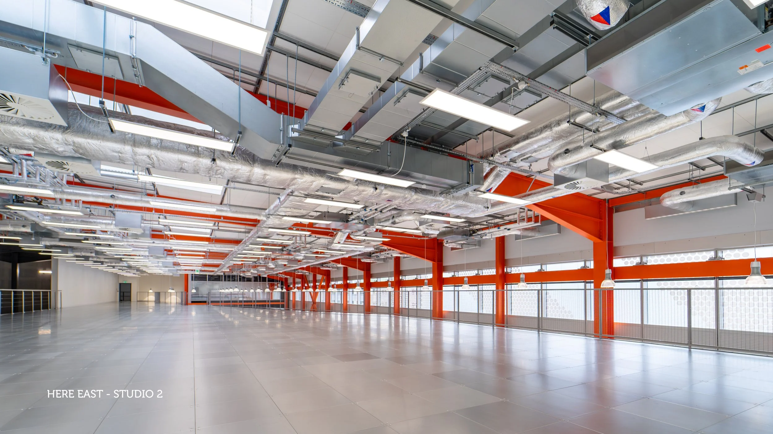 Empty, well-lit commercial space with white walls, gray tiled floor, and exposed ceiling with pipes and ventilation system.