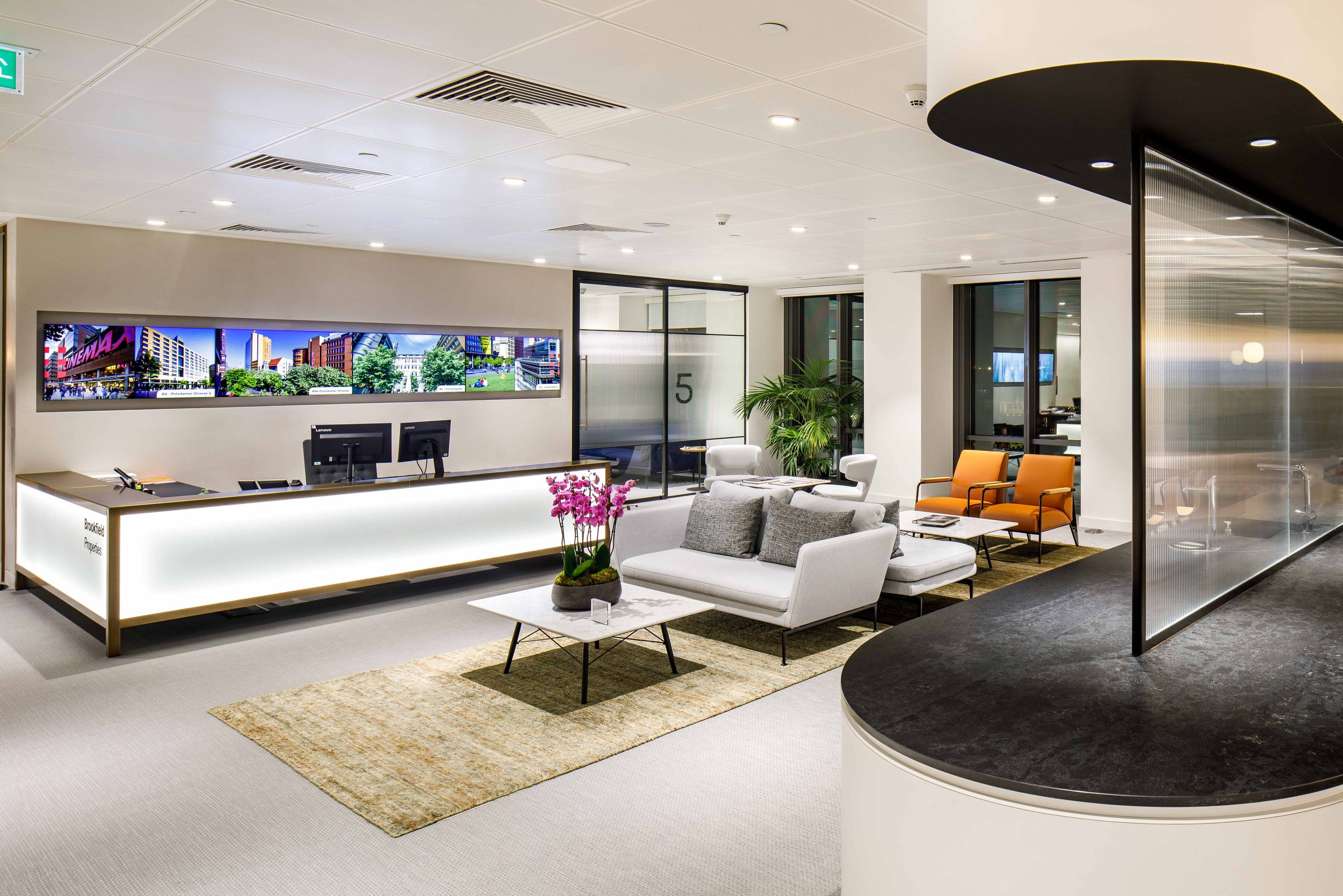 Modern office reception area with a desk, seating area with white and orange chairs, a large plant, and a digital display screen on the wall.
