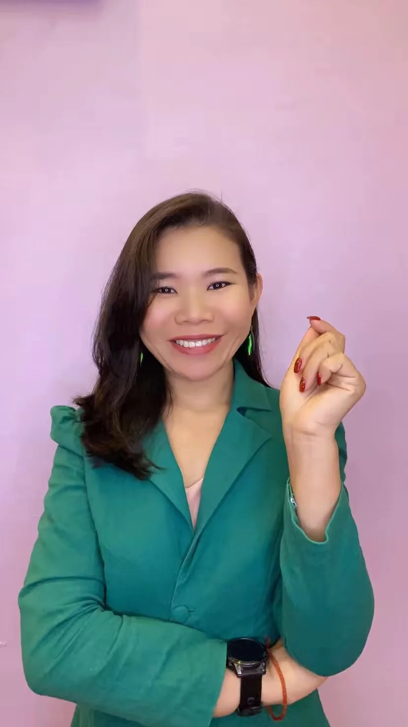 Smiling woman with shoulder-length dark hair, wearing a green blazer, green earrings, watch, and red nail polish, standing against a light pink wall.