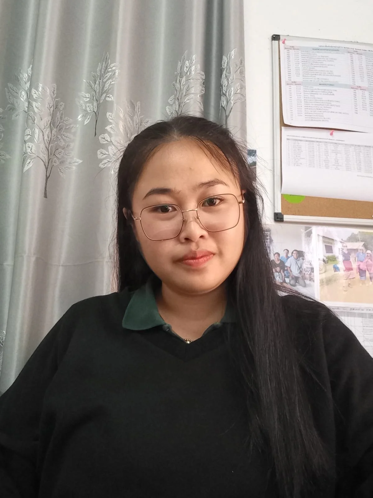 A young woman with long black hair, glasses, and a black sweater sitting in front of curtains and a bulletin board with papers and photos on the wall.