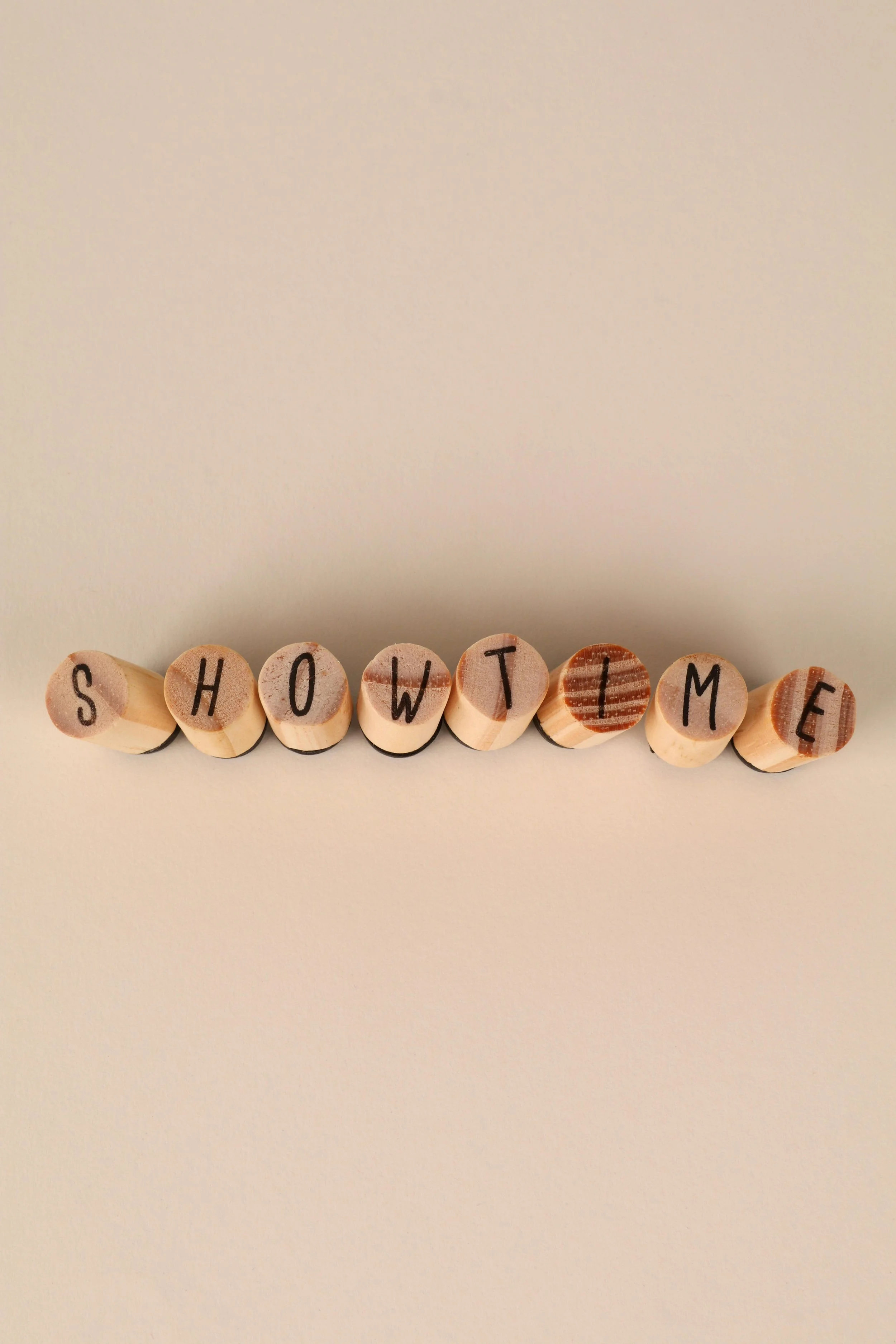Buchstaben auf kleinen runden Holzscheiben, die das Wort 'SHOWTIME' bilden, vor einem neutralen Hintergrund.