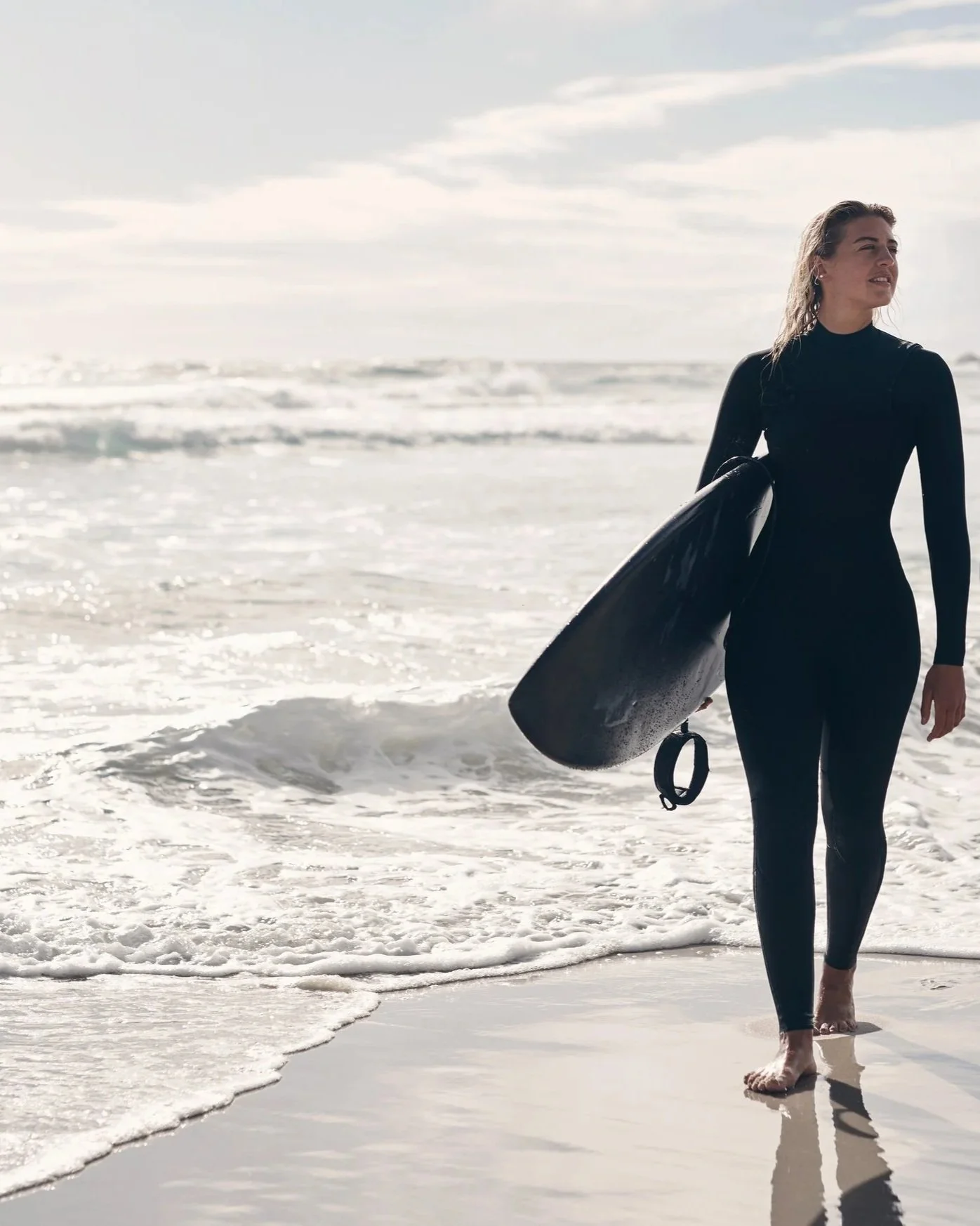 Frau in schwarzem Neoprenanzug mit Surfbretter am Strand, Meer und Himmel im Hintergrund.
