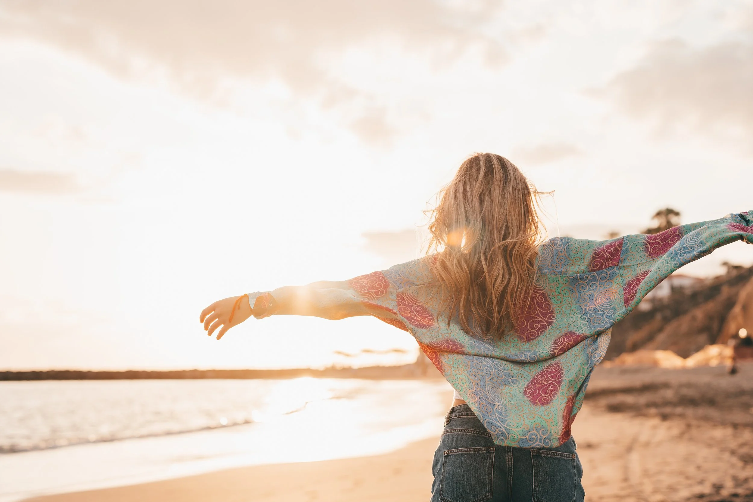 Eine Frau mit langen, lockigen Haaren, die an einem Strand im Sonnenuntergang mit ausgestreckten Armen steht.