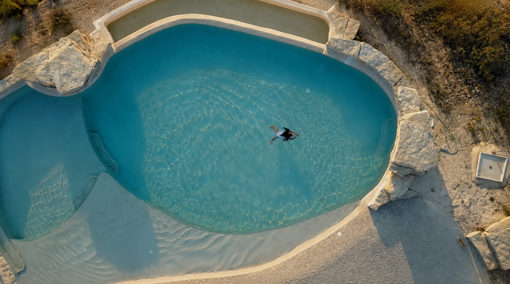 Ein Schwimmer liegt im leeren blauen Pool, umgeben von felsigen und sandigen Flächen, bei Sonnenuntergang.
