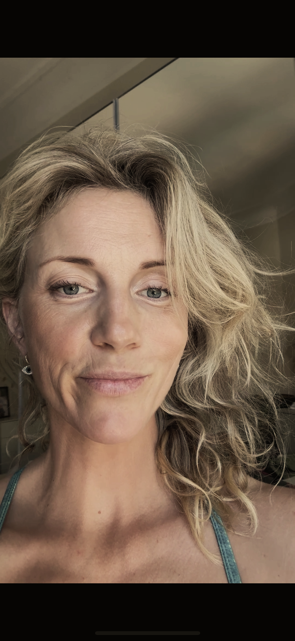 Nahaufnahme eines lächelnden Frauengesichts mit lockigem blondem Haar, hellen Augen und leichter Make-up im Innenraum.