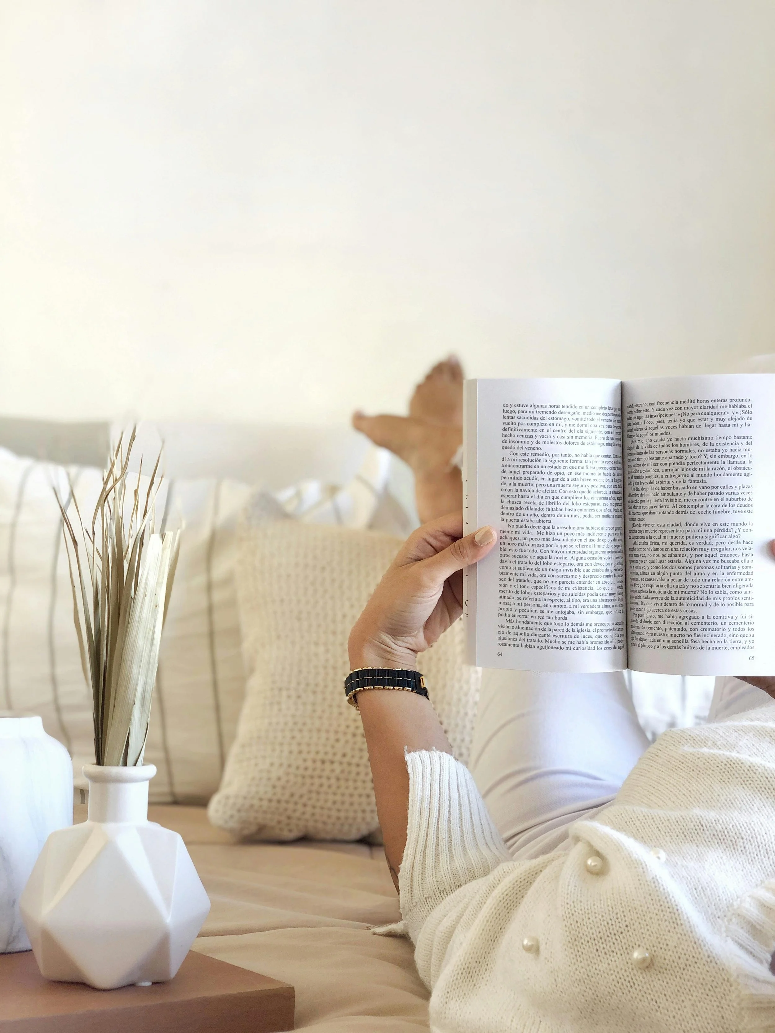 Eine Person liest ein Buch in einem hellen Raum, während sie auf einem Sofa liegt. Vor ihr auf dem Tisch stehen weiße Vasen mit getrockneten Zweigen.