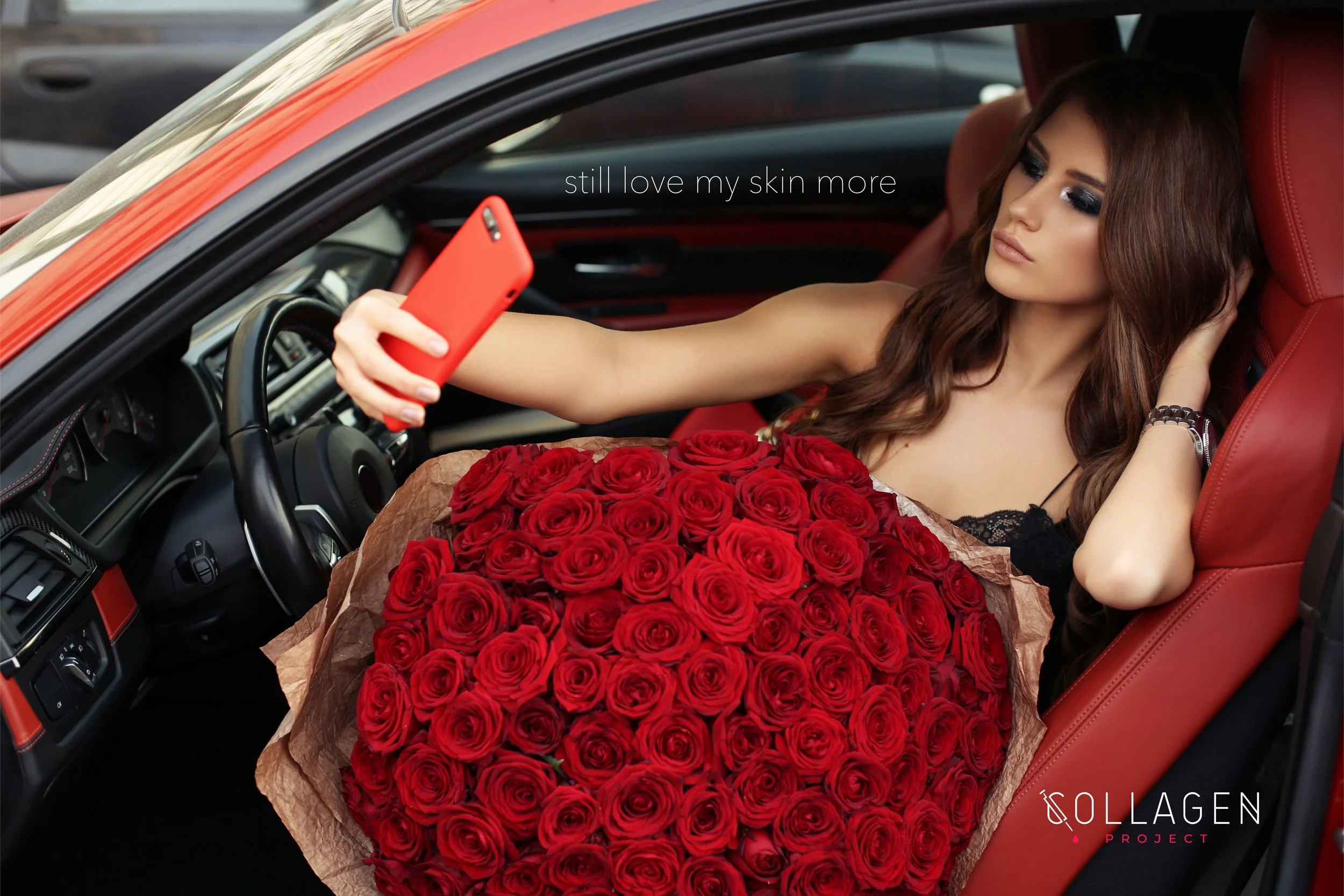 A woman with long brown hair and makeup is sitting in the driver's seat of a red car, wearing a black lace top, holding a large bouquet of red roses on her lap, and taking a selfie with an orange smartphone. The car has red seats and a black dashboar