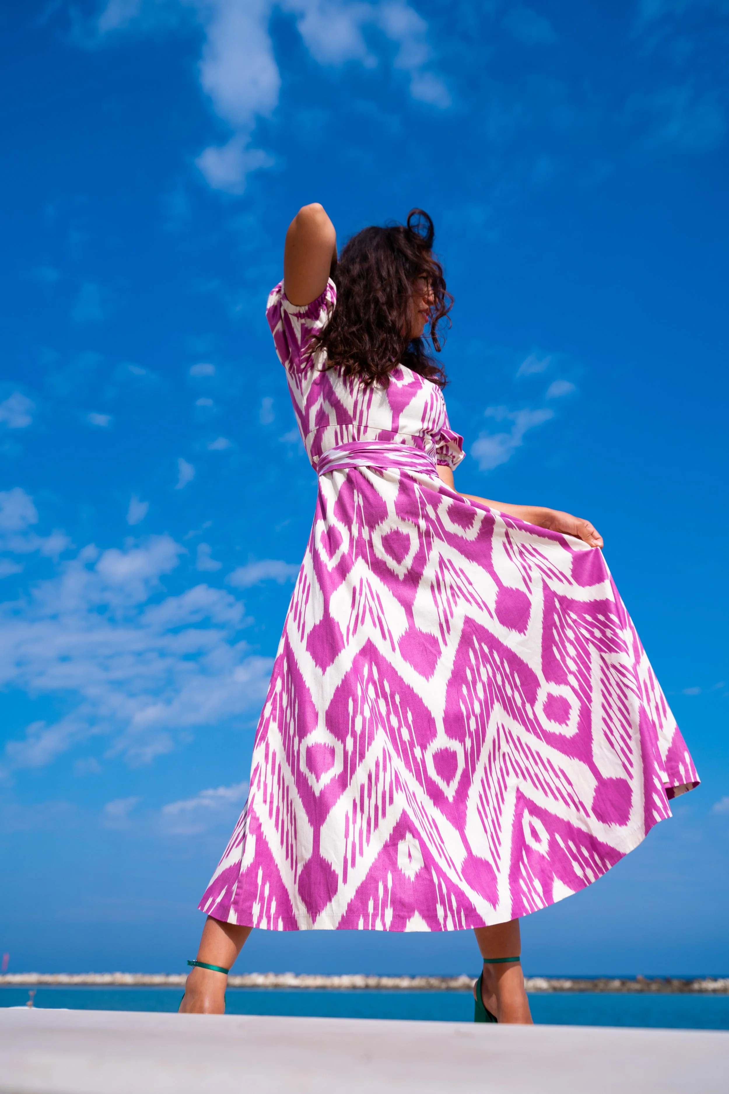Frau in einem pink-weißen Kleid steht vor blauem Himmel am Wasser.