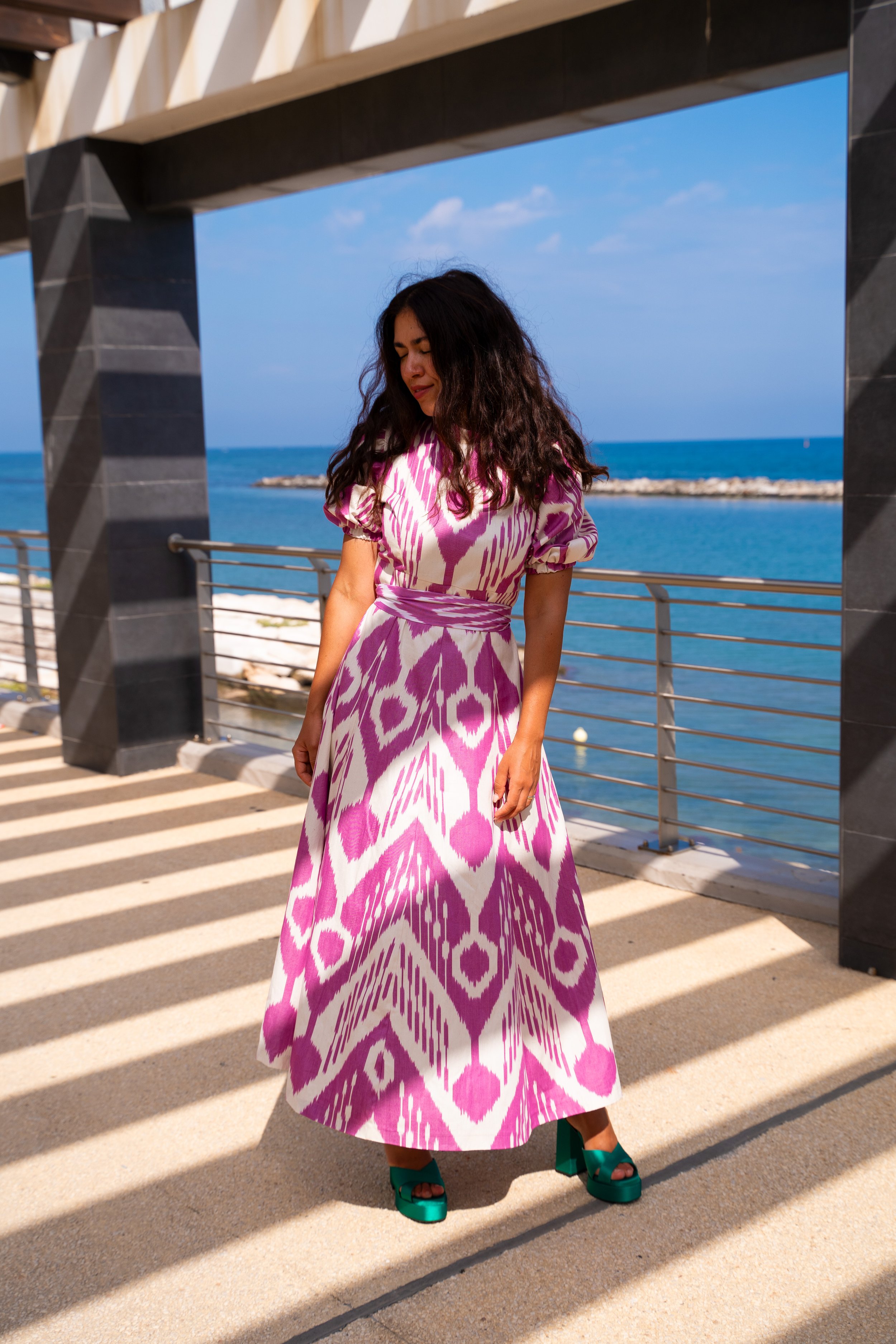 Frau in einem langen pink-weißen Kleid mit Muster, trägt grüne Schuhe, steht an einer Meerespromenade im Sonnenschein, Blick nach unten, im Hintergrund blauer Himmel und Meer.