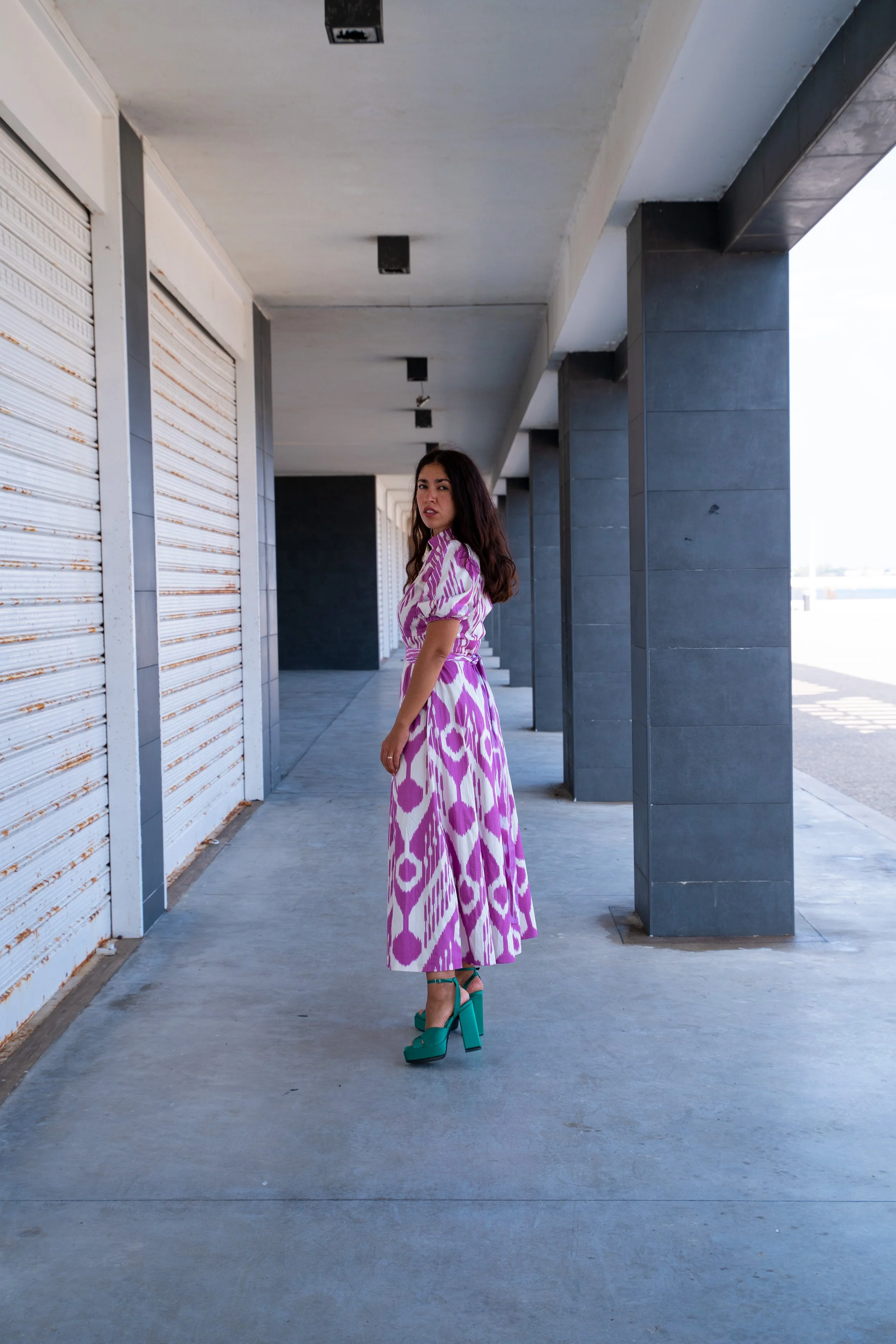 Eine Frau in einem langärmeligen, pink-weißen Kleid mit Leopardenmuster und hohen, grünen Sandalen steht in einer modernen, offenen Ladenzeile. Die Umgebung besteht aus grauen Wänden und einer hohen Decke mit quadratischen Beleuchtungsöffnungen.