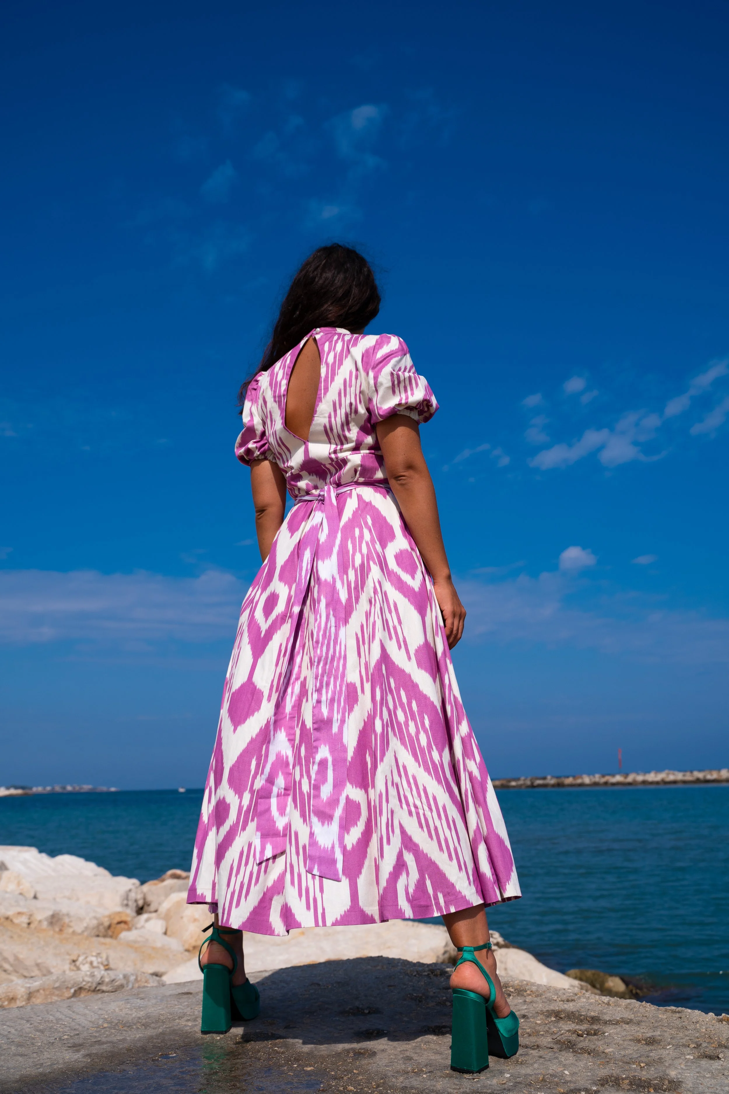 Frau in einem pink-weißen gestreiften Kleid mit tiefem Rückenausschnitt steht auf einer Steinterrasse am Wasser bei blauem Himmel.
