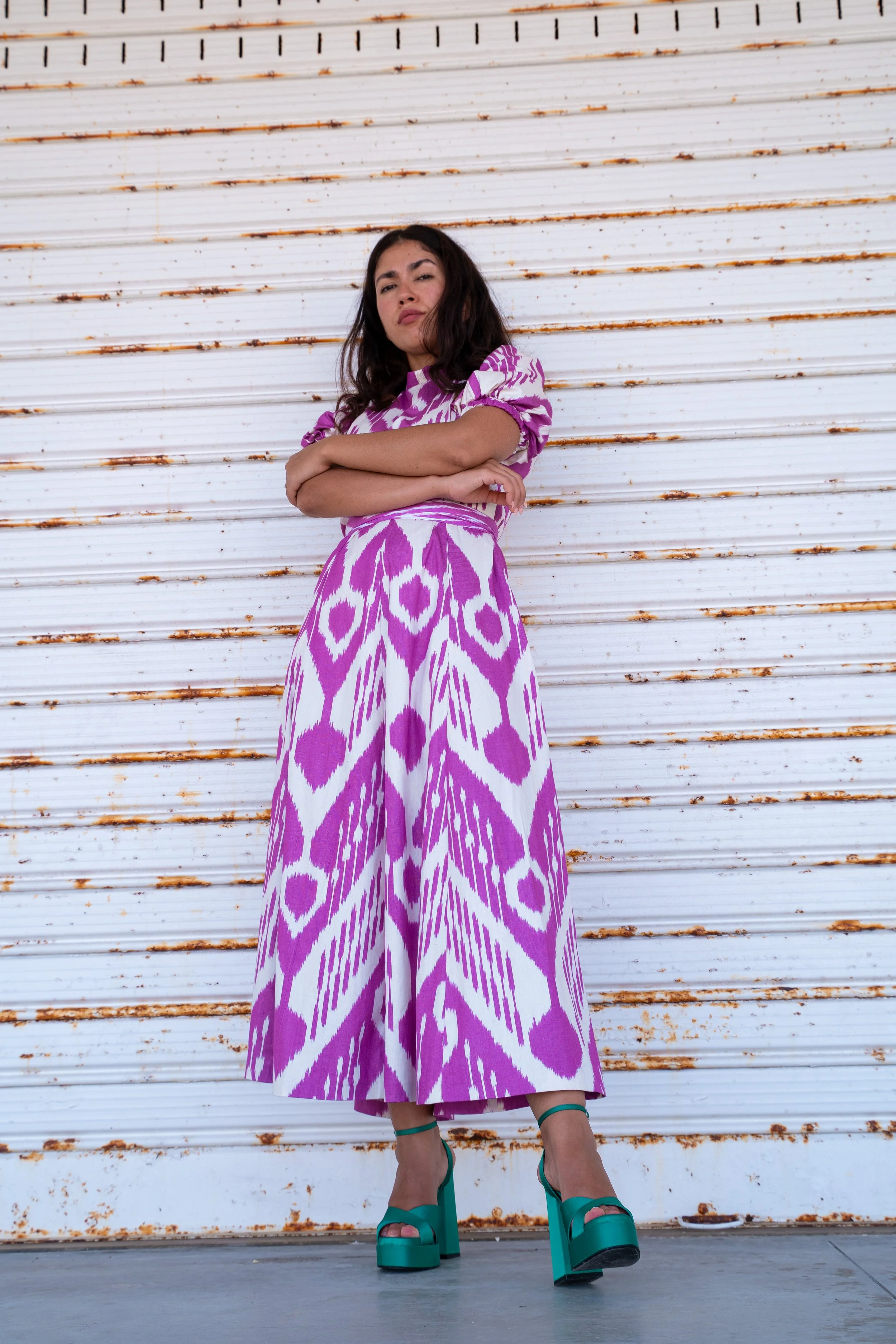 Junge Frau trägt lange, violett-weiße gemusterte Robe und hohe grüne Schuhe vor einer rostigen, weißen Metalltür.