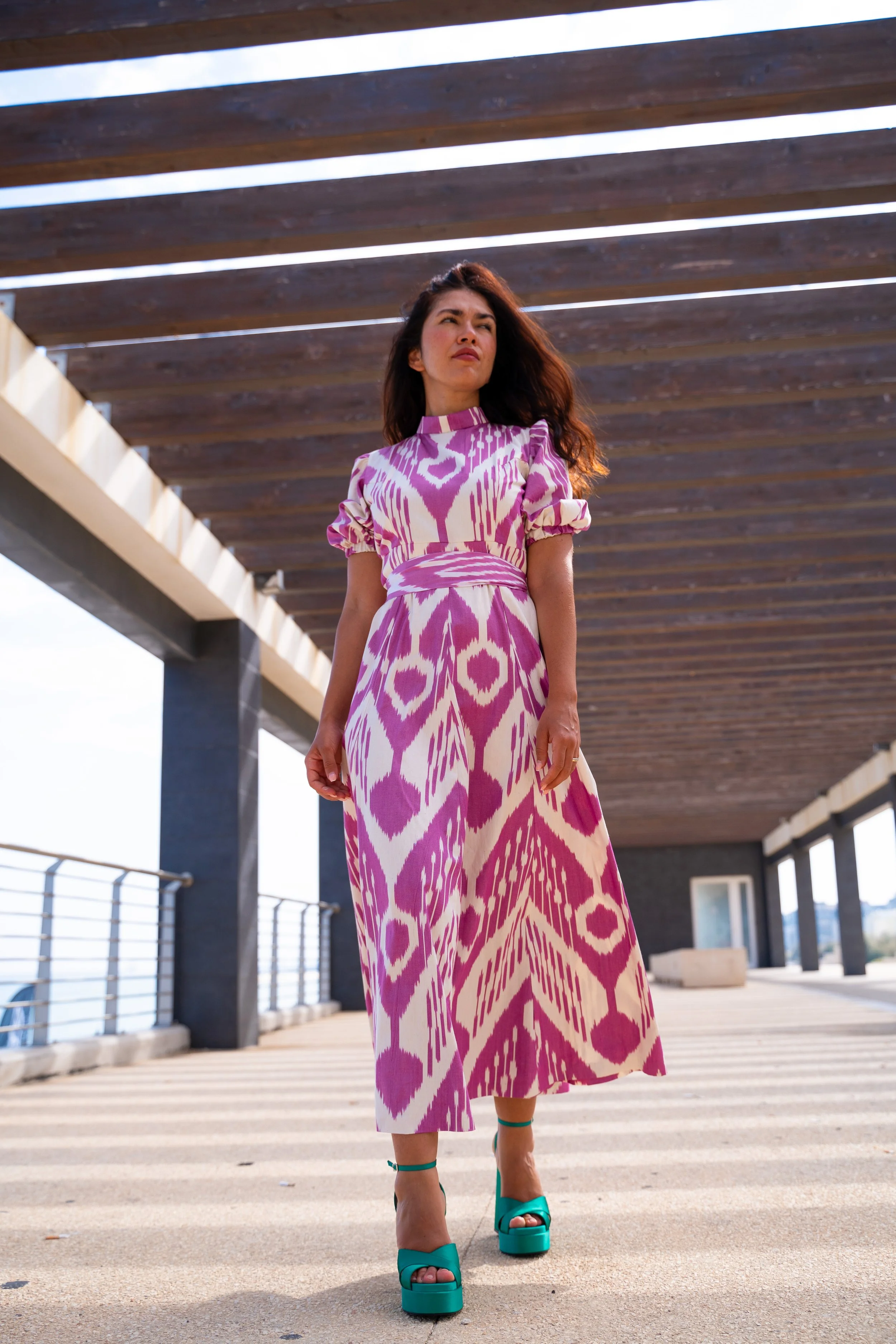 Frau in pink-weiß gemustertem Kleid und grünen High Heels steht unter einer modernen Holzüberdachung.