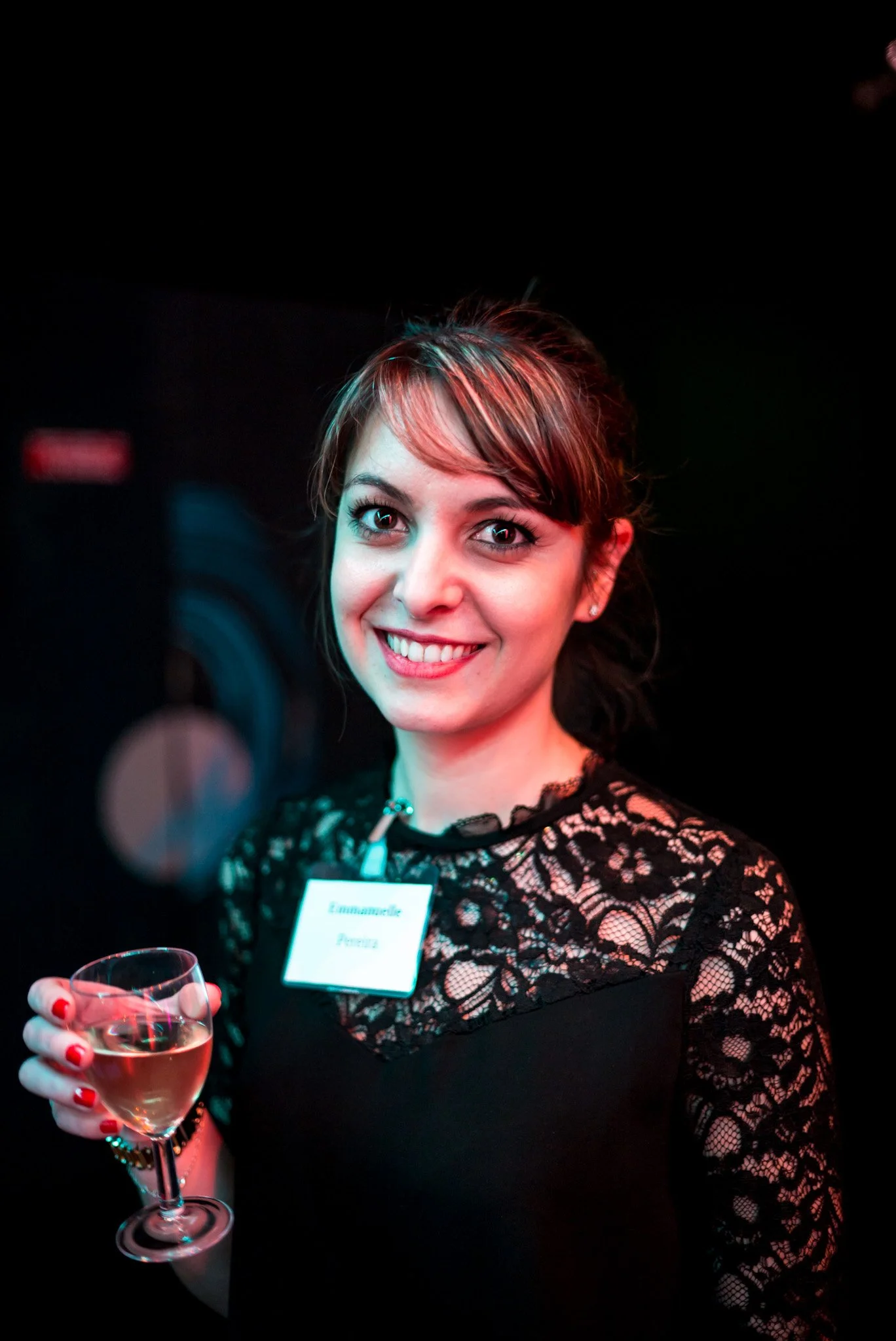 Emmanuelle Pereira sourit à l'objectif avec un verre de vin lors de la soirée Régus Paris. Robe noire en dentelle, ambiance sophistiquée et lumineuse, représentant l'élégance et la joie de l'événement.