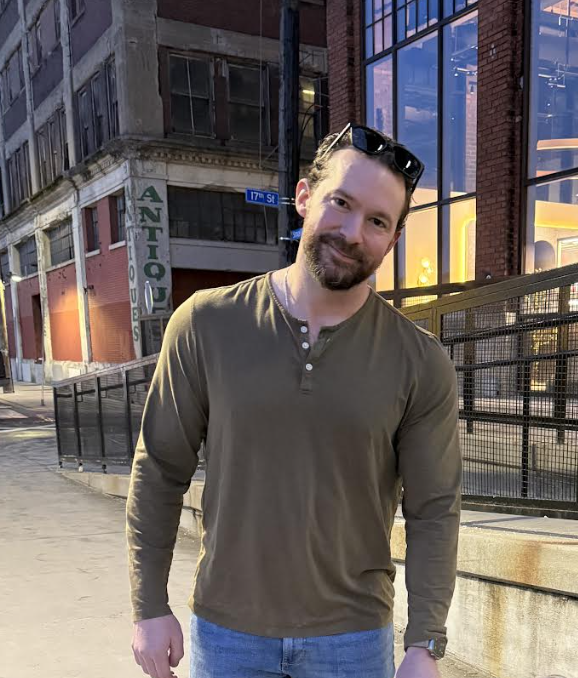 A man with a beard and sunglasses on his head, wearing a brown long-sleeve shirt and jeans, standing outdoors in an urban setting at dusk, with buildings and a lit window in the background.