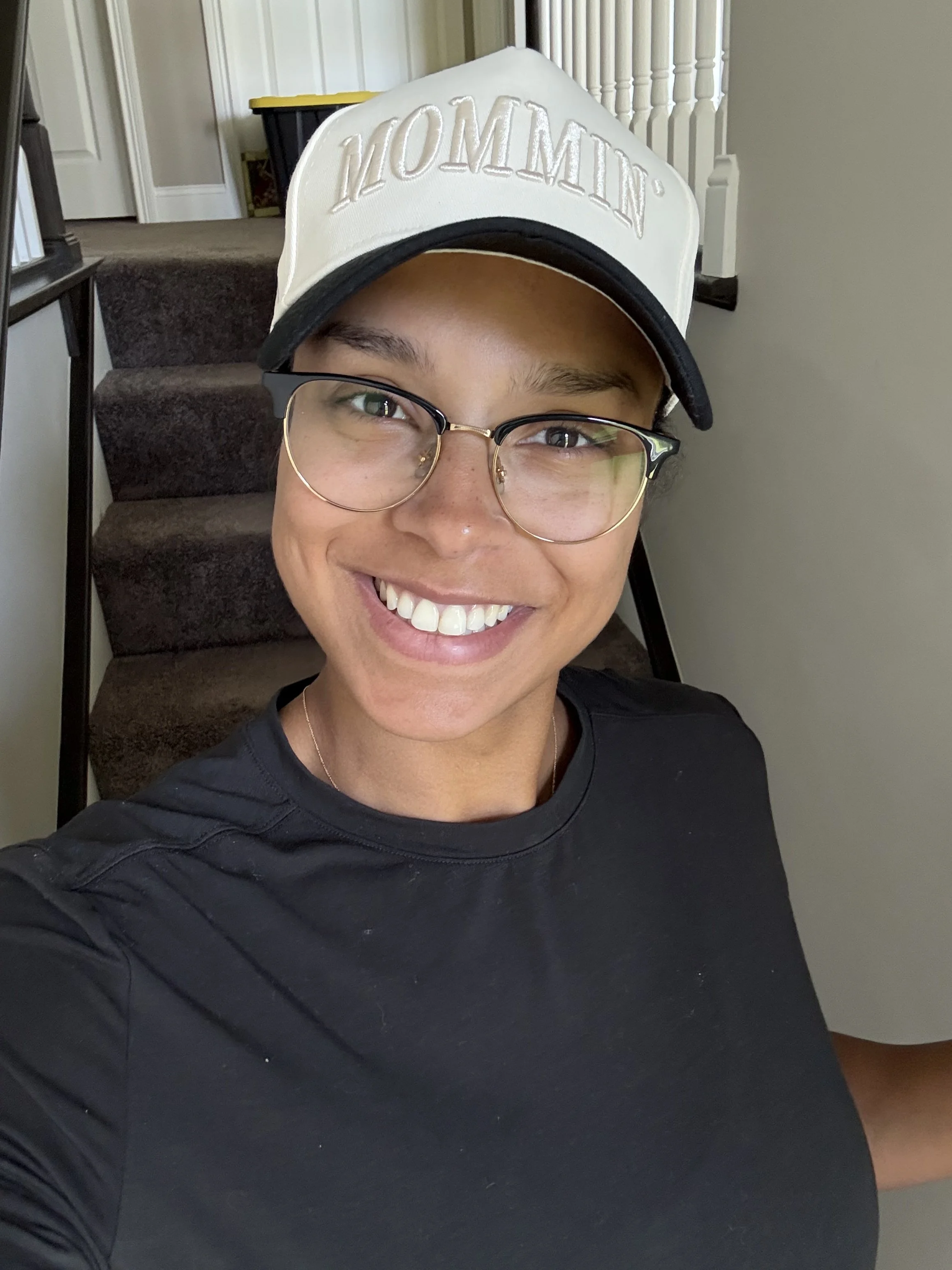 Person smiling wearing glasses, a white and black cap, and a black top, standing in front of stairs in a house.