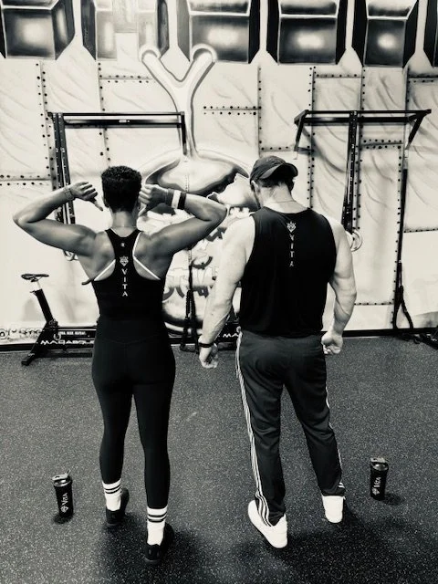 Two people standing in a gym with their backs to the camera, facing a wall with gym equipment and a large anatomical drawing of a human torso. They are wearing workout clothes, one with a tank top and the other in a sleeveless shirt. Water bottles are on the floor nearby.