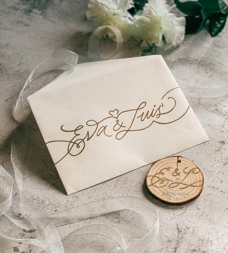 Una invitación de boda con los nombres 'Eva & Luis' escritos en letra cursiva dorada, ubicada en una superficie de color gris, acompañada de una flor blanca y una pieza de madera grabada con las iniciales 'E & L' realizada por Bendita Caligrafía.