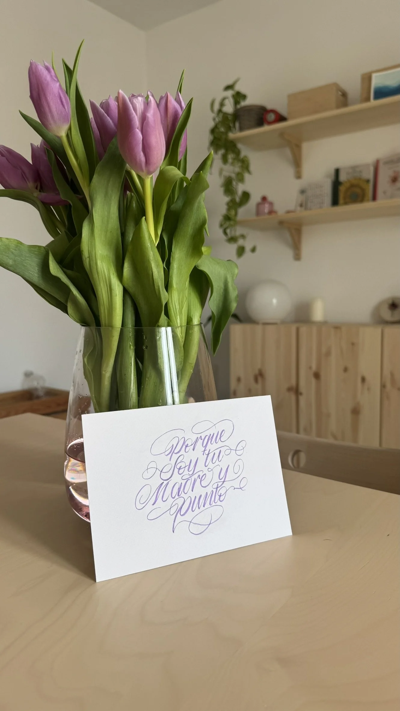 Un ramo de tulipanes rosas en un florero de vidrio en una mesa de madera, con una tarjeta que dice 'Porque soy tu madre y yo punto' hecho en una caligrafía por Bendita Caligrafía.