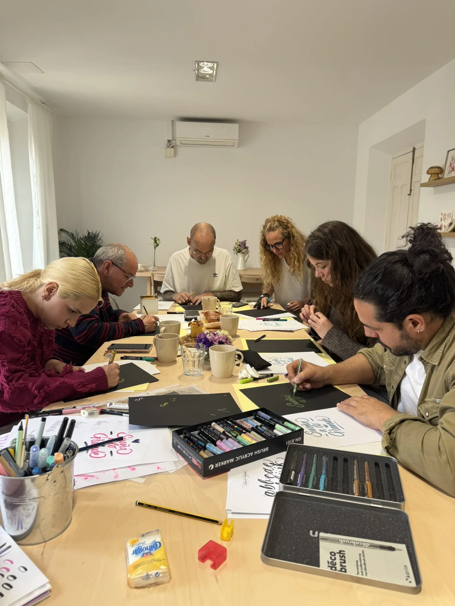 Varias personas sentadas alrededor de una mesa participando en una actividad de caligrafía y lettering organizada por Bendita Caligrafía, con marcadores, cuadernos y papel en la mesa.