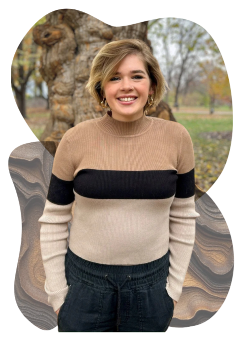 Young woman with short blonde hair smiling, standing outdoors near a large tree with autumn leaves.